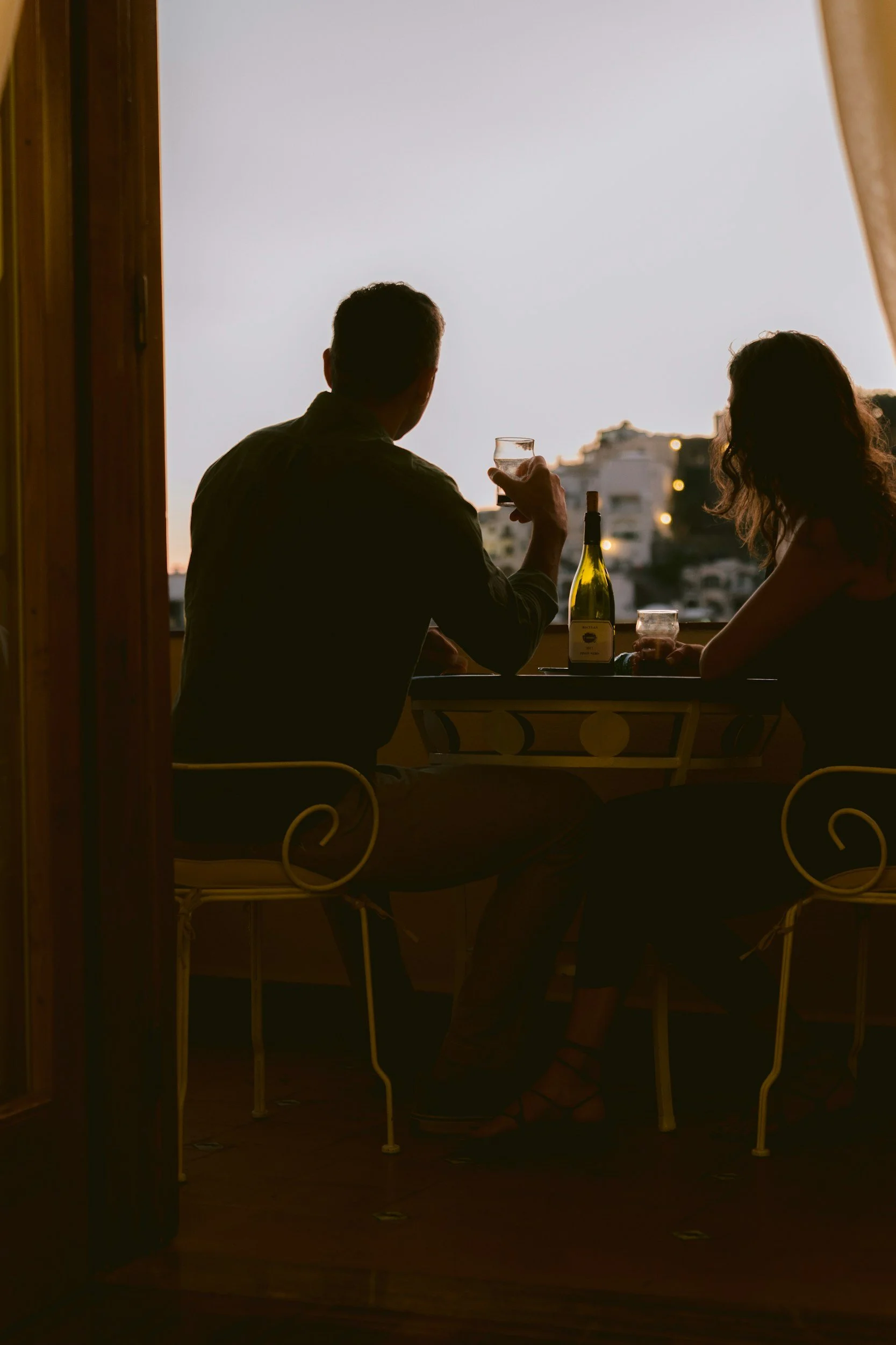 A man and woman sitting at a table, facing a large window with a cityscape view at sunset. The man holds a glass and there is a bottle on the table.
