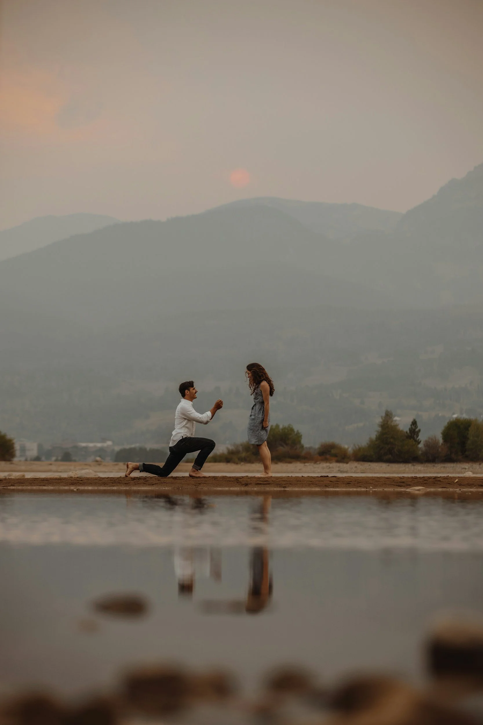 A man kneeling and holding a woman's hand at the edge of a reflective body of water during sunset with mountains in the background.