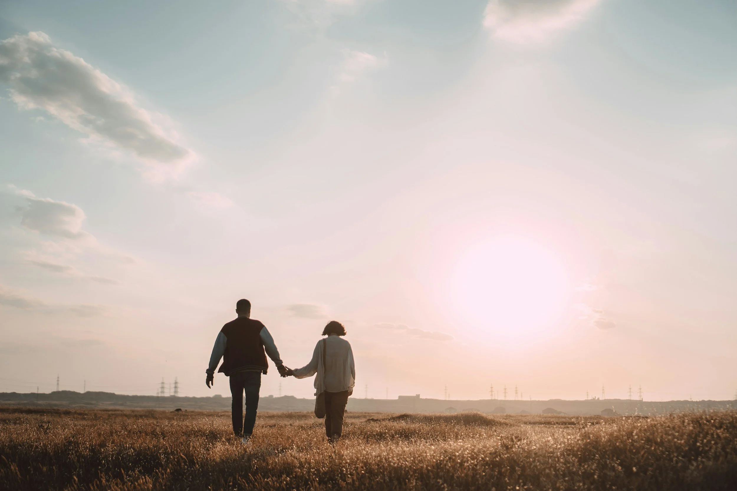 A couple holding hands walking through an open field during sunset with a few clouds in the sky.