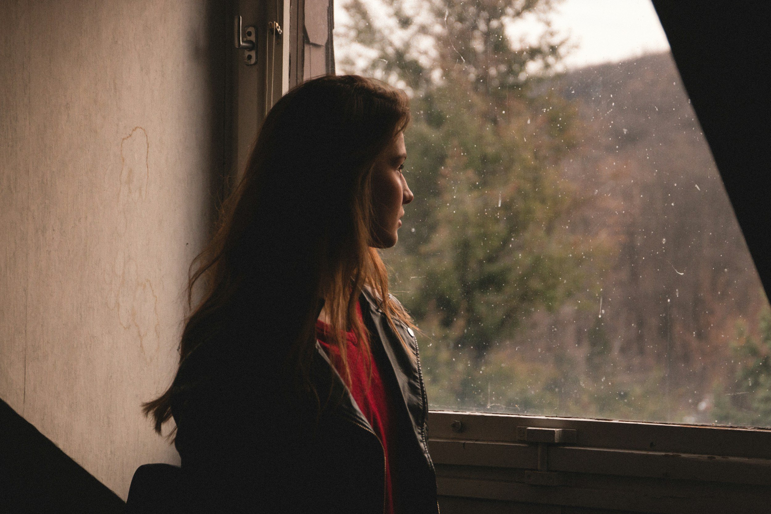 A woman with long brown hair, wearing a black jacket, standing by a window on a rainy day, looking outside at blurred trees and raindrops on the glass.