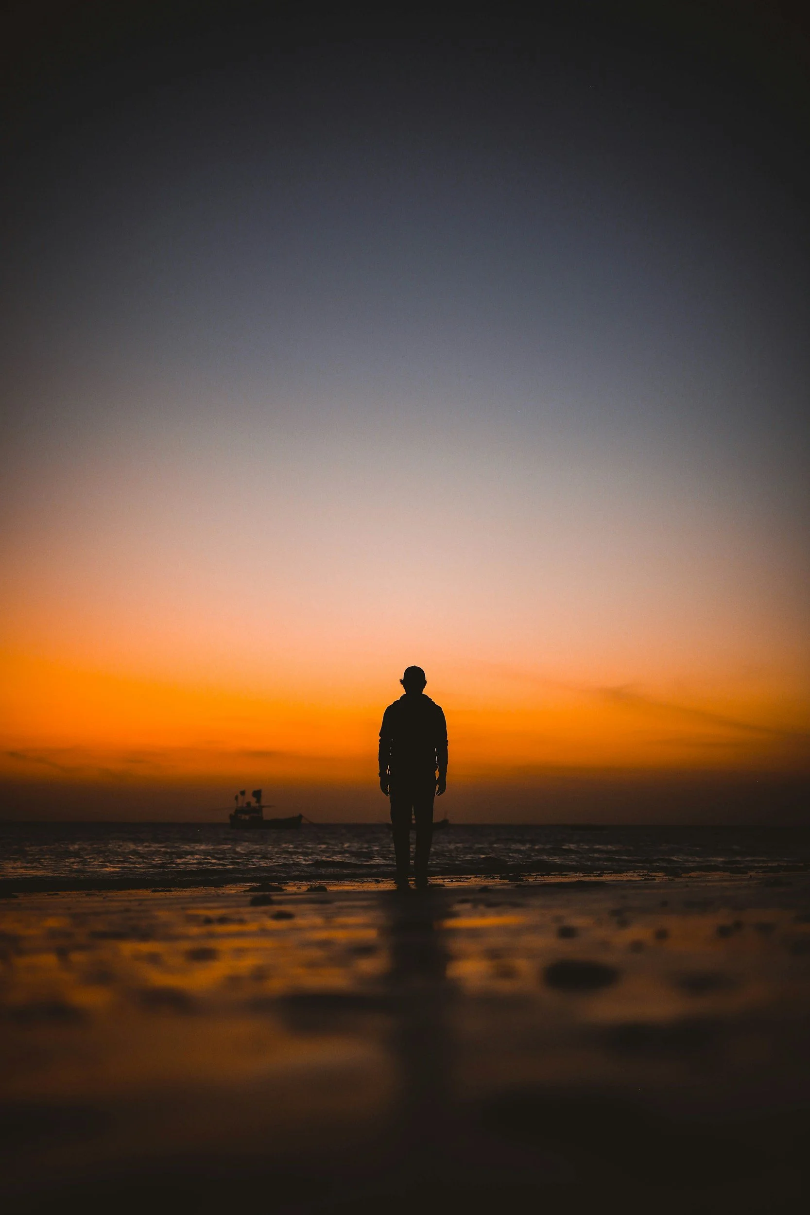 Person standing on the beach during sunset with a boat in the water and a colorful sky.