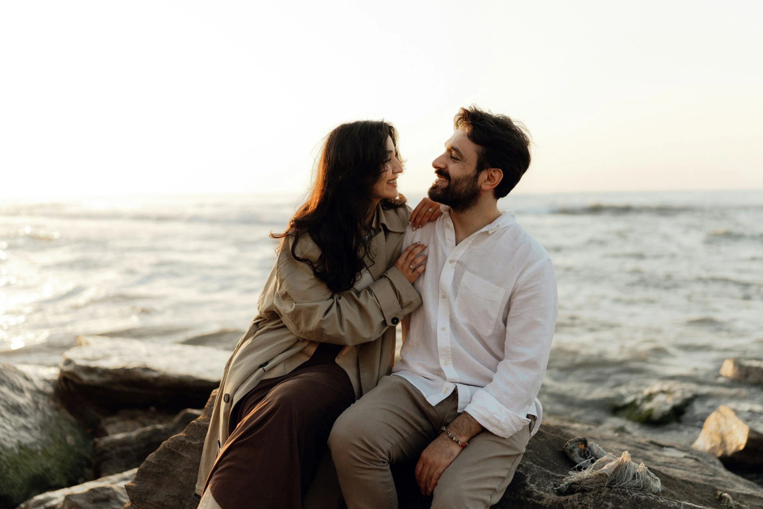 A couple sitting on rocks by the ocean, smiling and looking at each other as the sun sets.