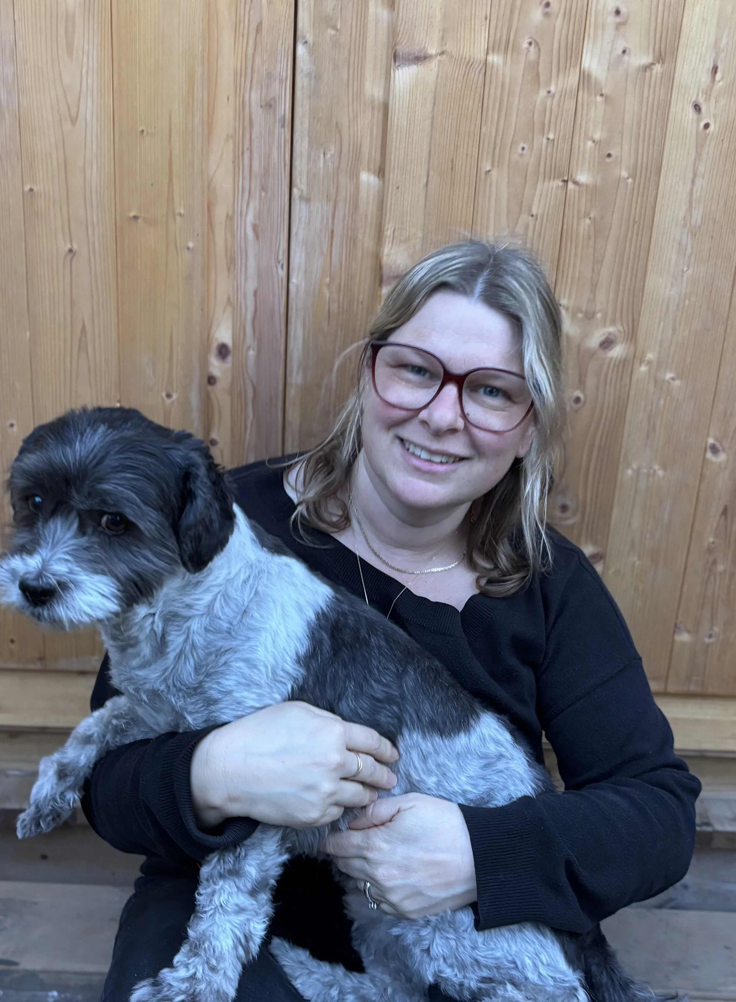 A therapist with blond hair and glasses smiling while holding a small black and white puppy, against a wooden paneled background.