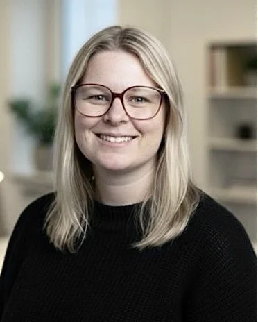 Portrait of a young woman with blonde hair, glasses, and a black sweater, smiling indoors with shelves in the background.