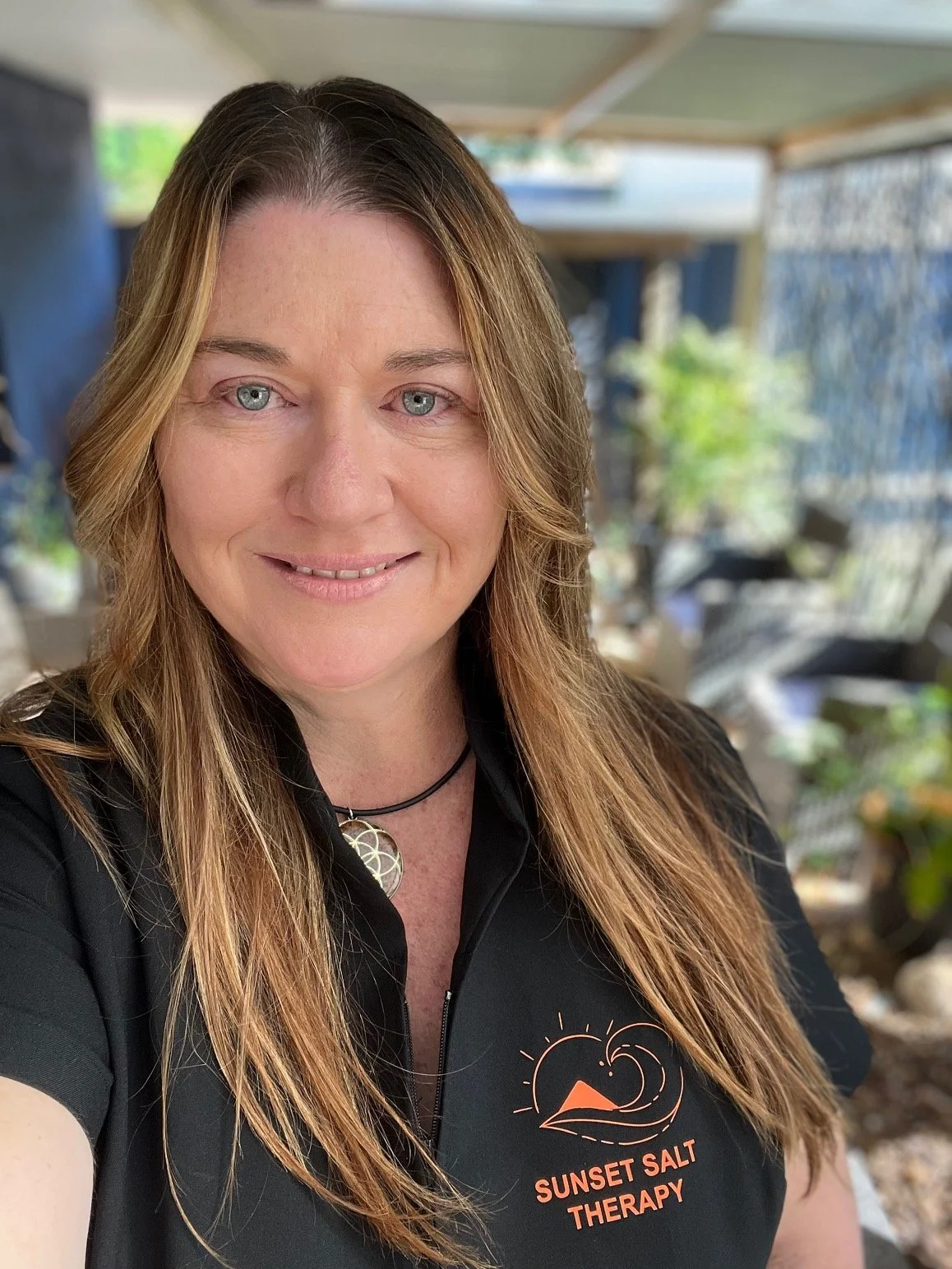 A woman with long, light brown hair and blue eyes smiling at the camera, wearing a black shirt with a sunset and ocean wave design and the words "Sunset Salt Therapy." She has a necklace with a round pendant, and the background appears to be an outdoor or patio setting with blurred greenery and furniture.