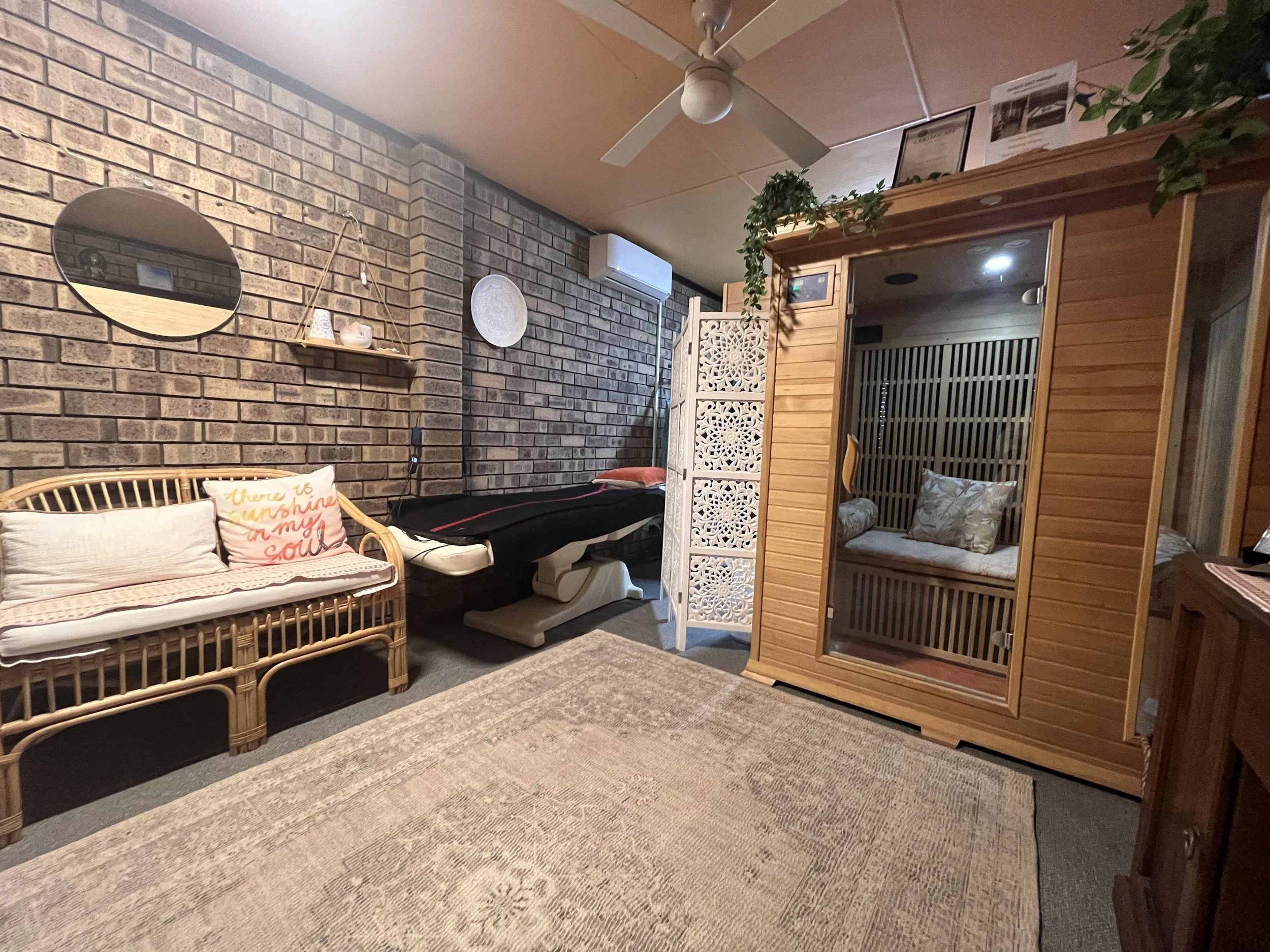 A room with a brick wall, a wicker bench with cushions, an ironing board, a wooden sauna with glass door, a folding white decorative screen, and various wall decorations including a round mirror and a white plate. There is a ceiling fan and an air conditioning unit.