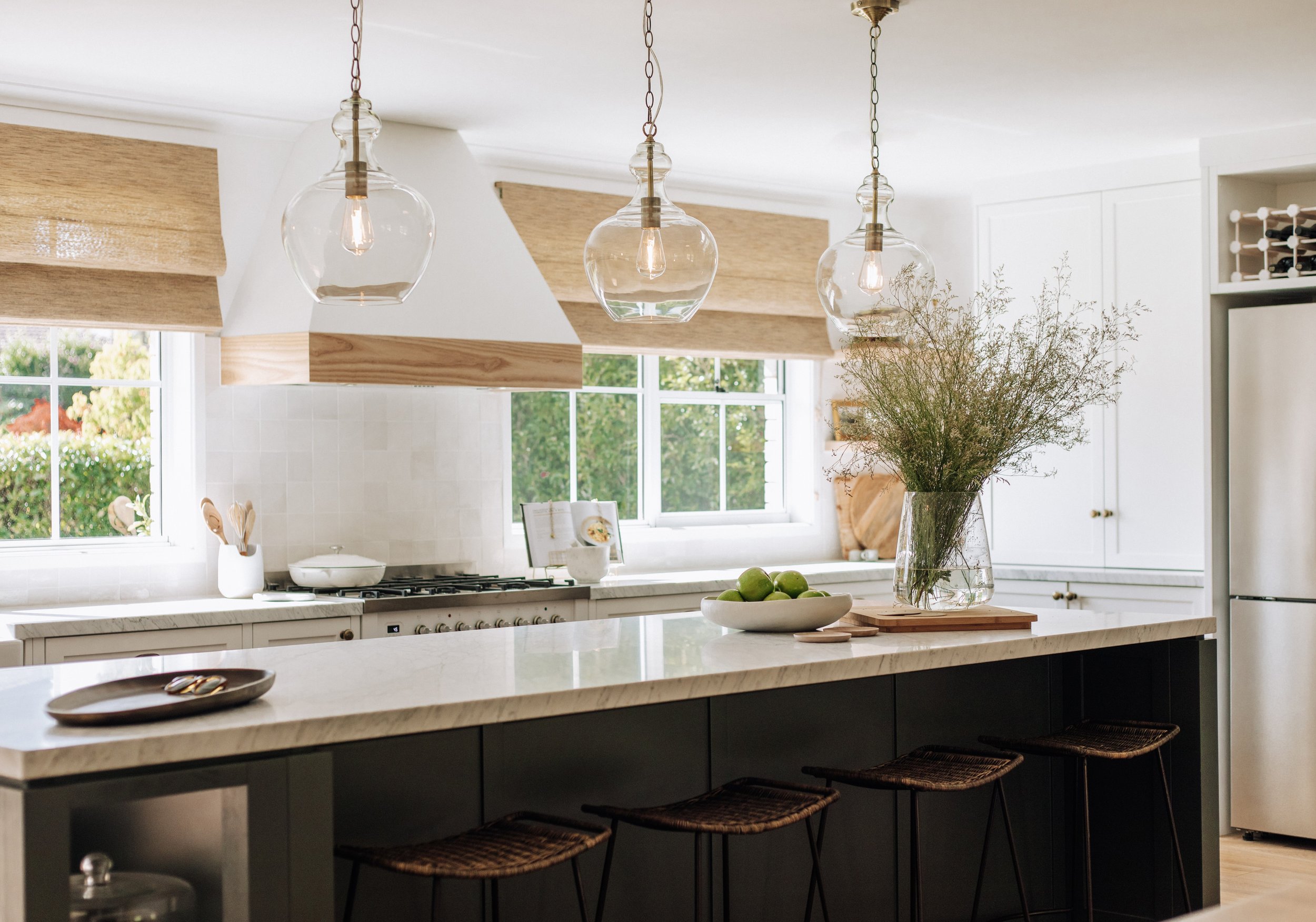 Modern kitchen designed by Oak & Olive Homes with white cabinets, marble countertops, dark island bench, bar stools, glass pendant lights, backed by large windows overlooking the garden