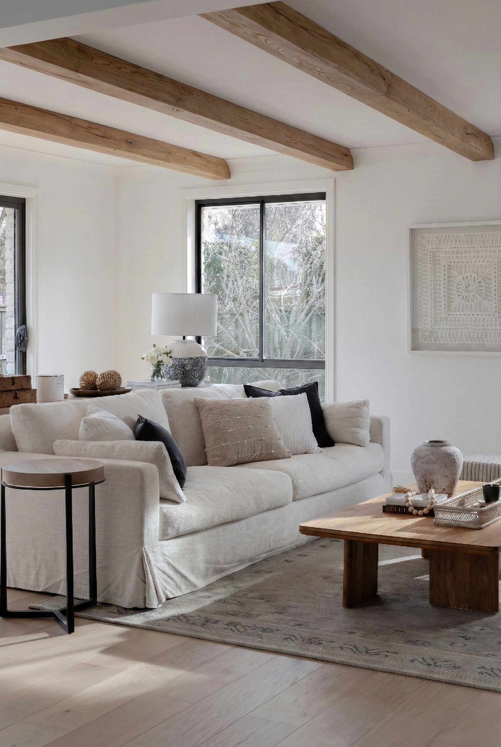 Bright, modern, cozy living room by Oak & Olive Homes featuring a large, off-white slipcovered sofa, wooden coffee table, light oak wood floors, and large windows overlooking winter trees