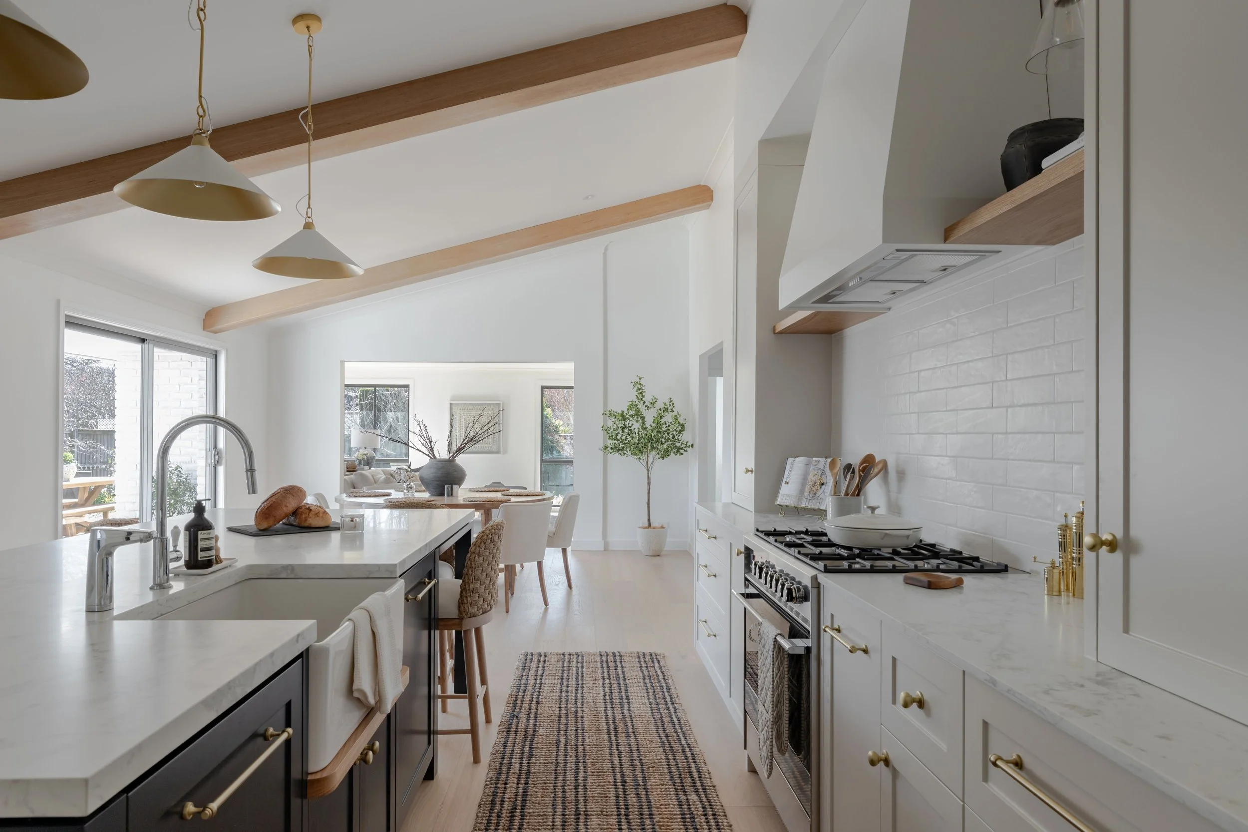Spacious kitchen by Oak & Olive Homes with white cabinetry, marble countertops, and a large island featuring a sink and woven barstools. Open layout leads to a dining area with light wood beams, natural decor, and sliding glass doors to the patio.
