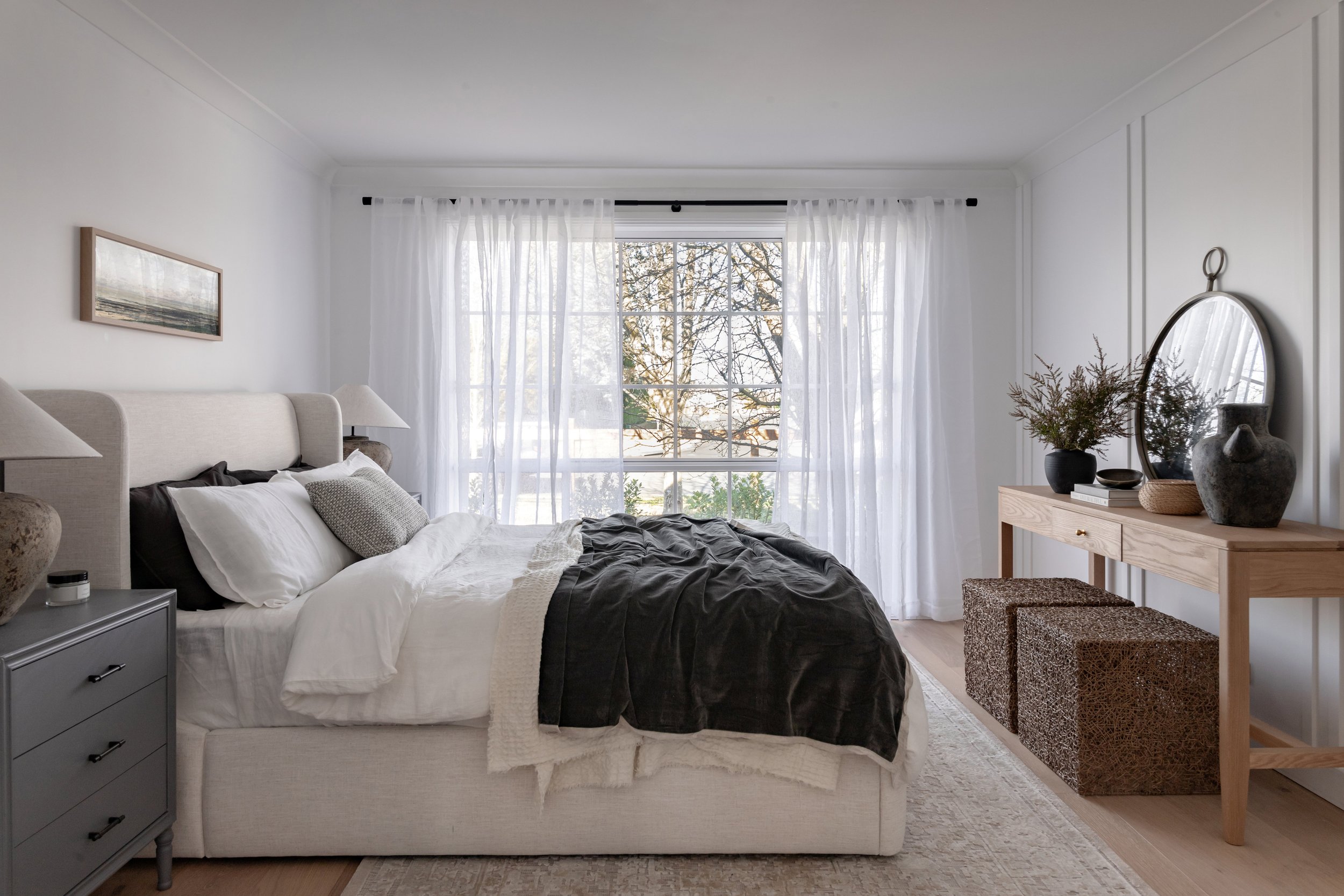 Master bedroom by Oak & Olive Homes with a neutral palette, upholstered bed, layered bedding, & sheer white curtains framing a large window. Features a wooden console table with decorative vases, round mirror, and textured ottomans for added warmth a