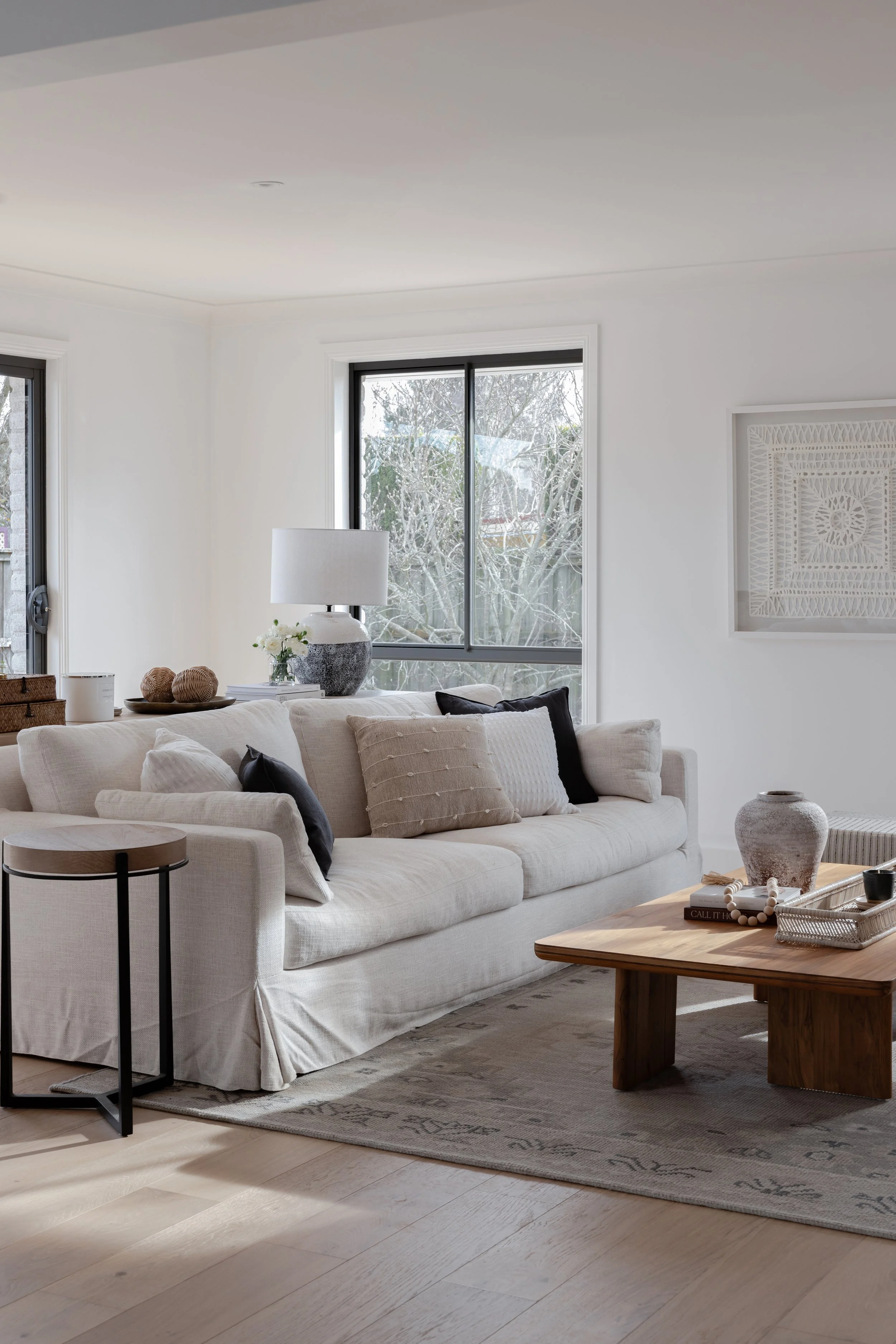 Bright, modern, cozy living room by Oak & Olive Homes featuring a large, off-white slipcovered sofa, wooden coffee table, light oak wood floors, and large windows overlooking winter trees