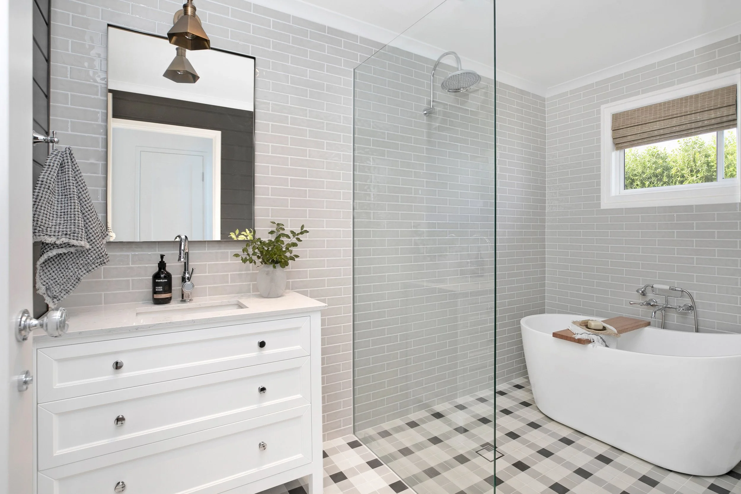 Country bathroom by Oak & Olive Homes featuring soft greige subway wall tiles, a white vanity, frameless glass shower and freestanding bathtub. Natural textures, chrome fittings and a woven roman blind add warmth to the light, classic space.