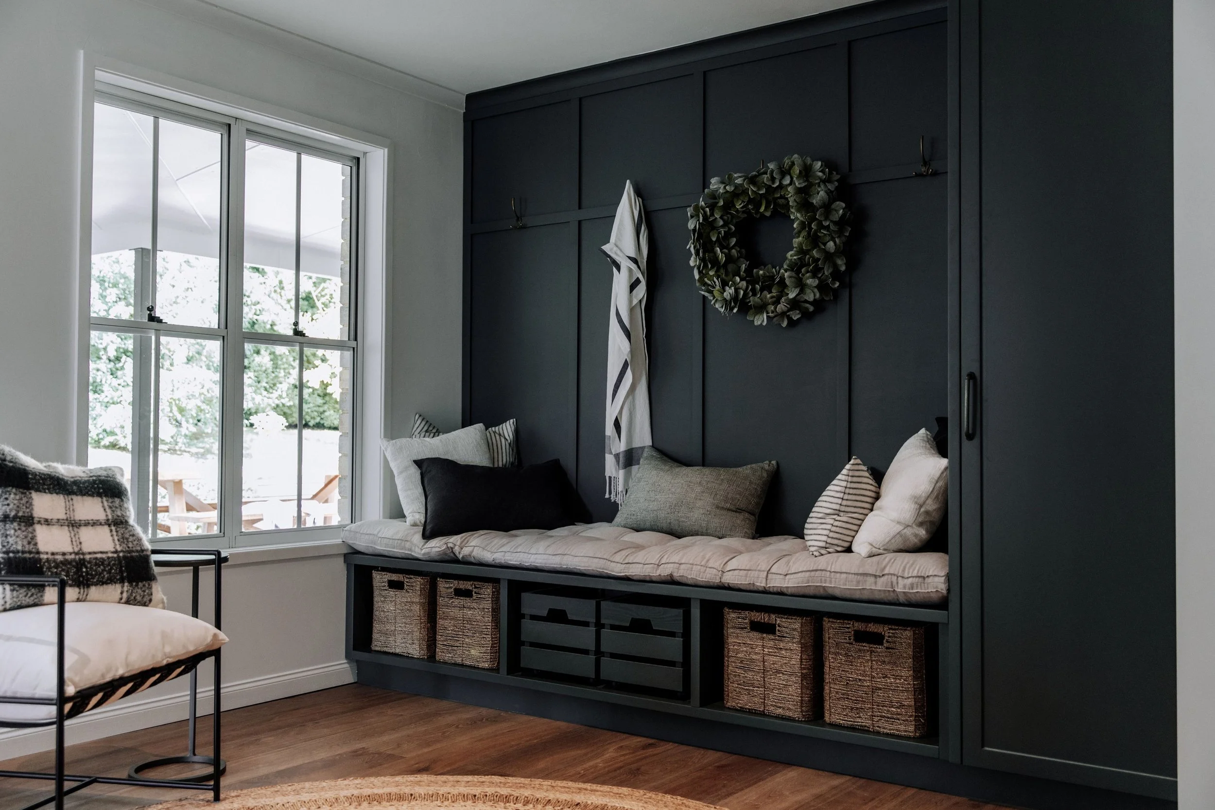 A dark grey green mudroom by Oak & Olive Homes with wainscotting on the wall and a cushioned bench with wicker baskets below for storage