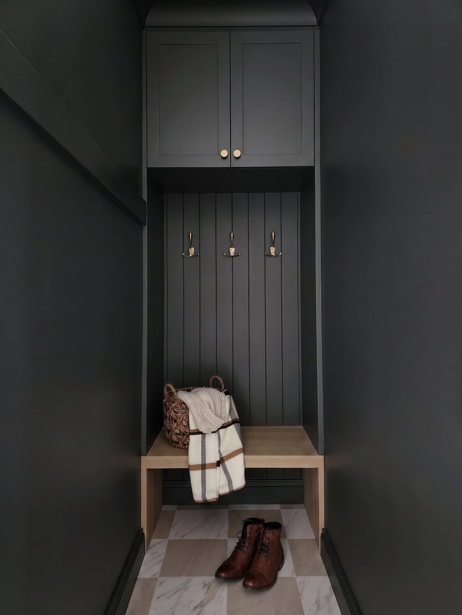 Dark charcoal mudroom nook designed by Oak & Olive Homes, featuring built-in cabinet storage, brass coat hooks, vertical wall paneling, and a natural wood bench. A woven basket, plaid scarf, knit throw, and brown leather boots add cozy entryway styli