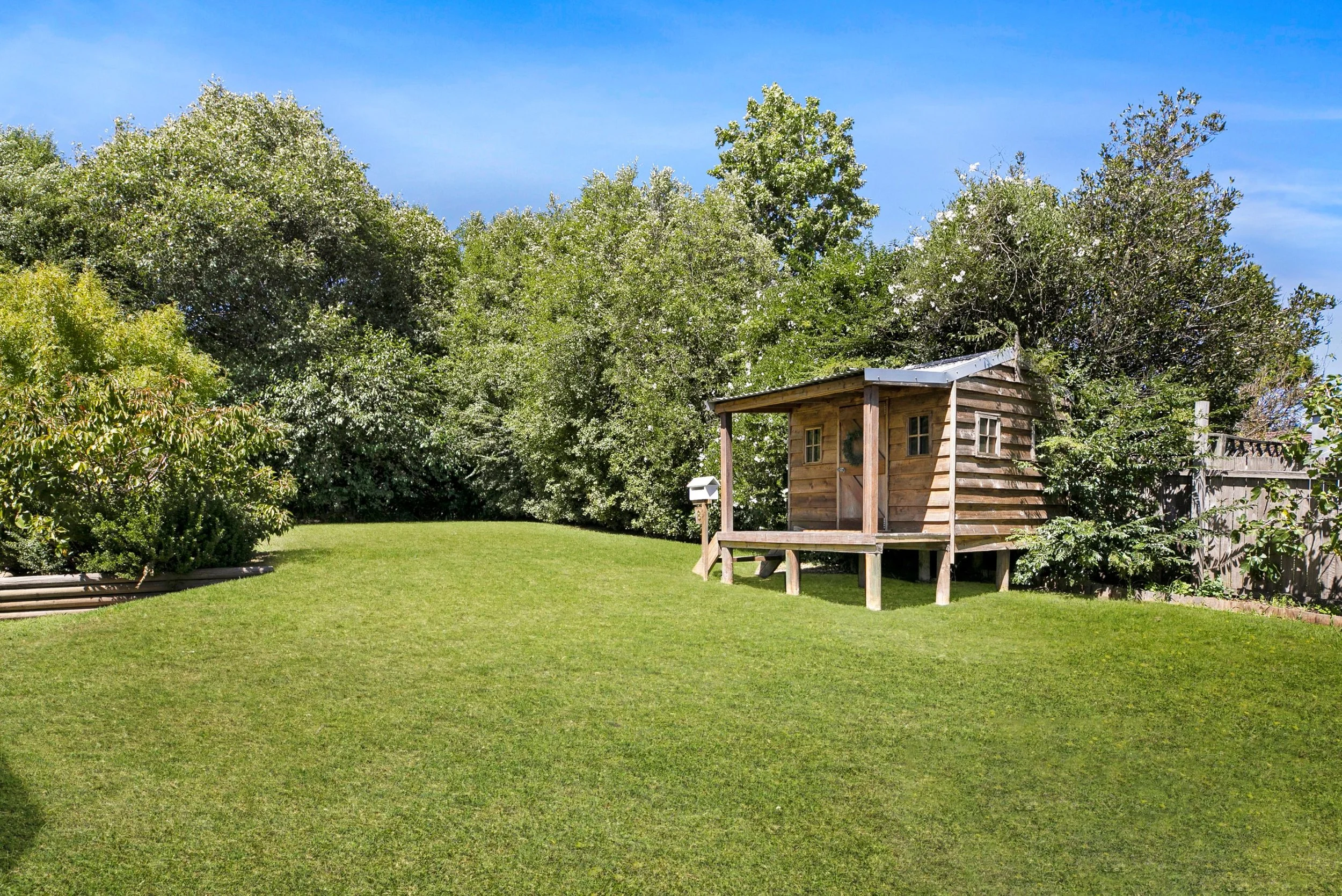 Generous sized backyard with luscious green grass and established trees for privacy. A log cubby house located amongst the trees
