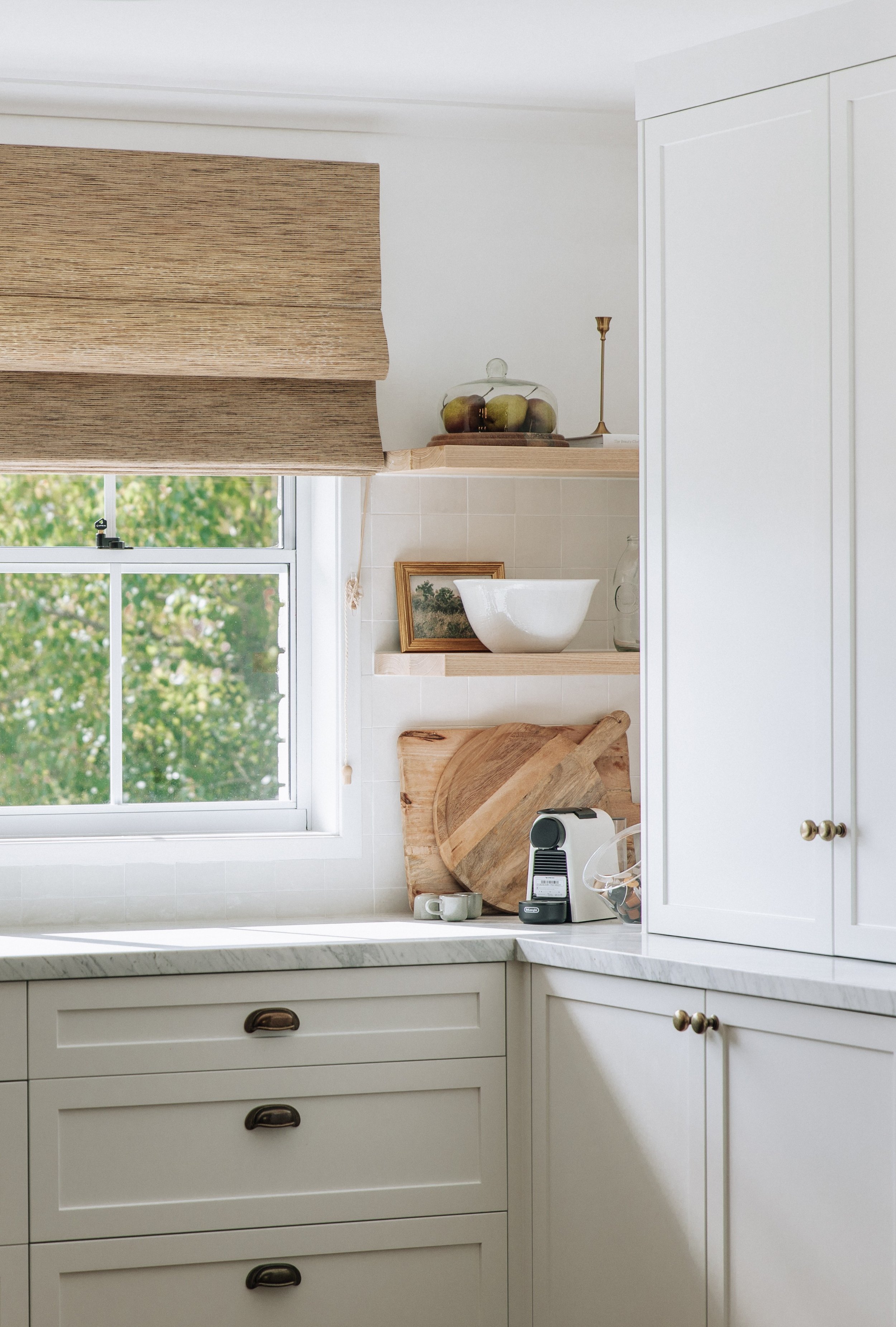 Charming kitchen corner by Oak & Olive Homes with cream shaker cabinets, marble countertops, and open wood shelves styled with ceramics, art, and a cloche of fruit. A window with a natural woven Roman shade brings in soft daylight above a compact esp
