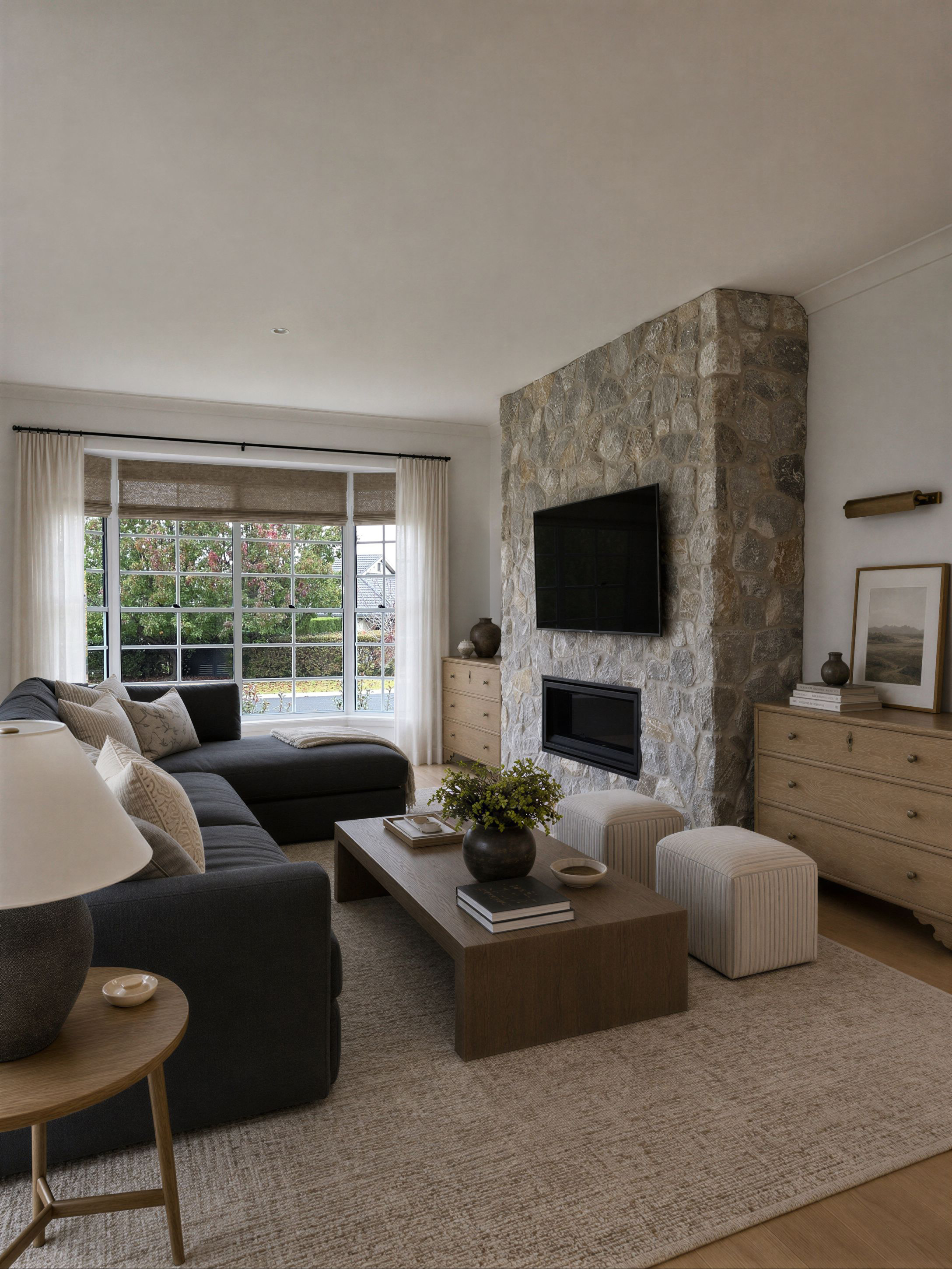 Oak & Olive Homes interior design featuring a cozy modern living room with a stone fireplace wall, mounted TV, dark sectional sofa, neutral linen curtains, natural wood furniture, and layered beige textures. Warm organic styling, soft contemporary de