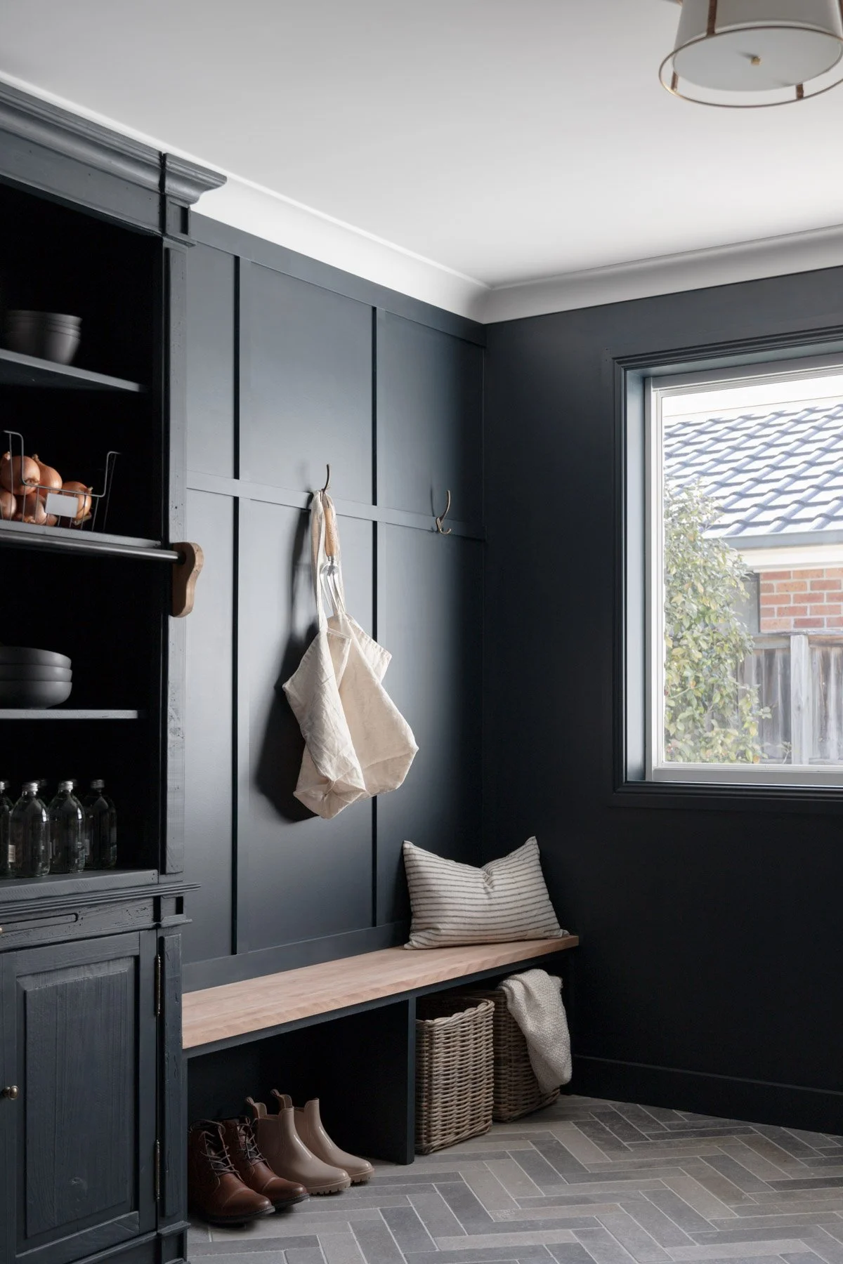 Stylish mudroom by Oak & Olive Homes with dark navy panel walls, wood bench seating, woven storage baskets, and neutral-toned boots arranged neatly below. Features a large window for natural light, wall hooks with canvas bags, and a cozy striped pill