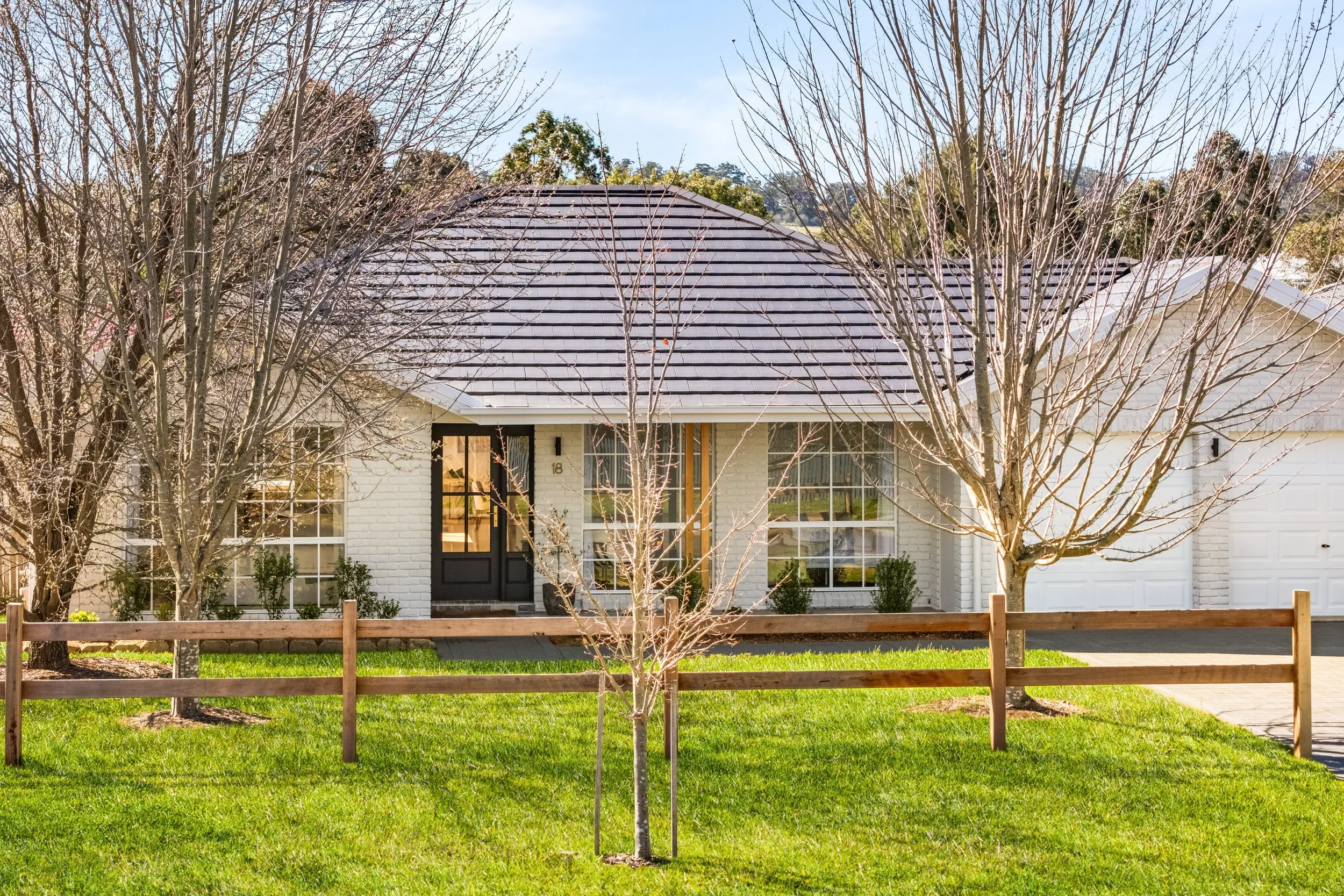 Charming single-story white brick house, designed by Oak & Olive, with a dark-trimmed front door, large windows, manicured lawn, wooden fence, and leafless trees in the front yard