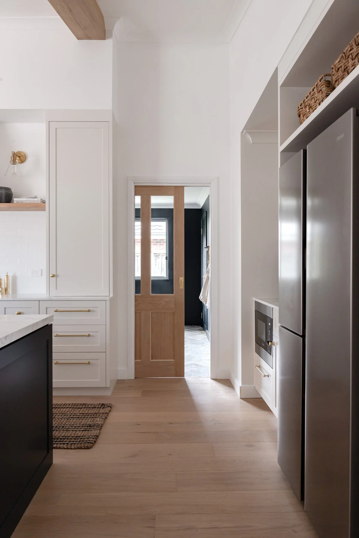 Kitchen hallway featuring light wood flooring, white cabinetry with brass hardware, and a stainless steel refrigerator. A sliding wooden door with glass panels opens to a dark-accented mudroom with herringbone stone tile flooring.
