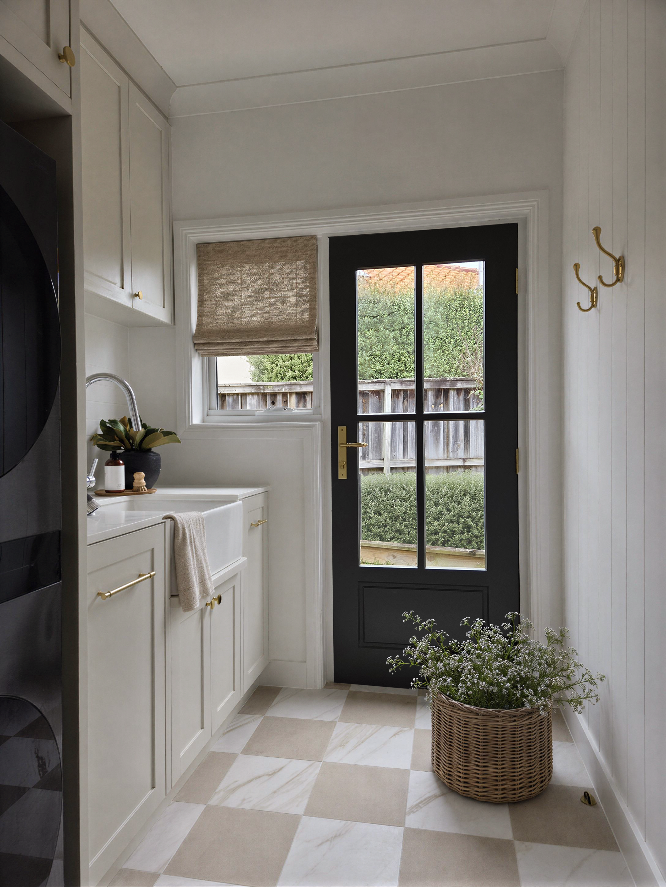 Oak & Olive Homes interior design featuring a refined laundry with cream cabinetry, brass hardware, charcoal French door, woven Roman shade, checkerboard tile floor, and natural wicker basket styling. This elegant transitional home design blends warm