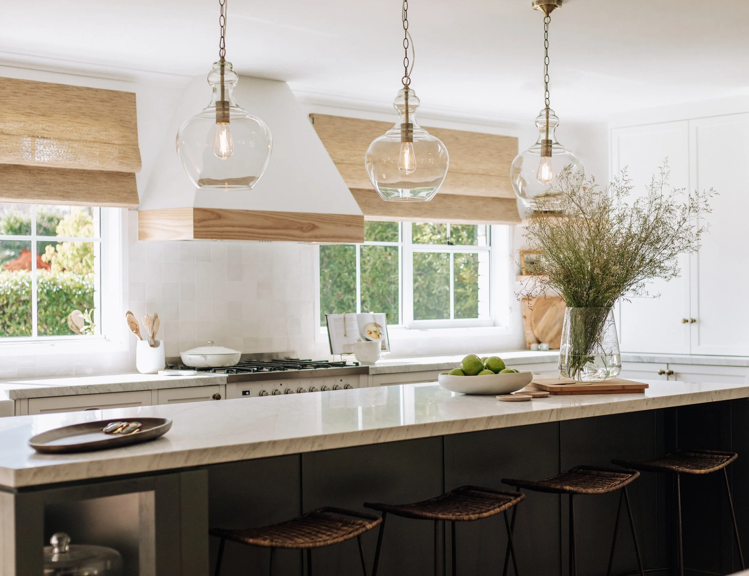 Modern kitchen designed by Oak & Olive Homes with white cabinets, marble countertops, dark island bench, bar stools, glass pendant lights, backed by large windows overlooking the garden