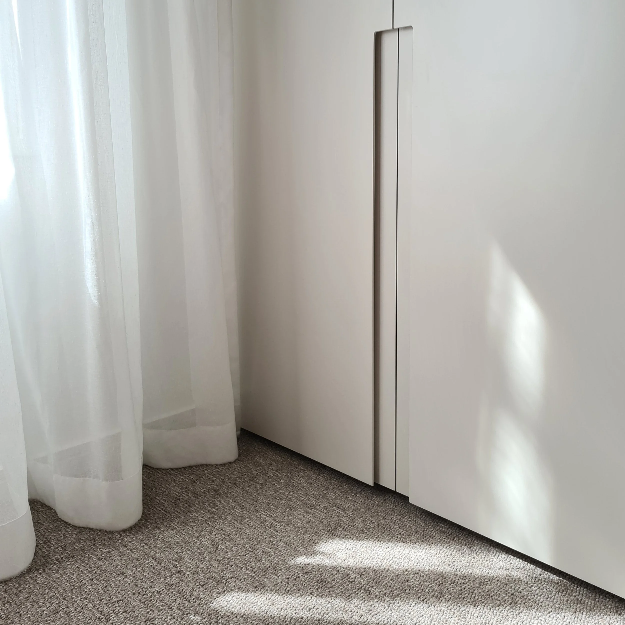 Minimalist interior corner featuring sheer white curtains, soft beige carpet, and a sleek modern wardrobe with integrated handle. Sunlight filters in gently, casting natural shadows for a calm, serene ambiance.
