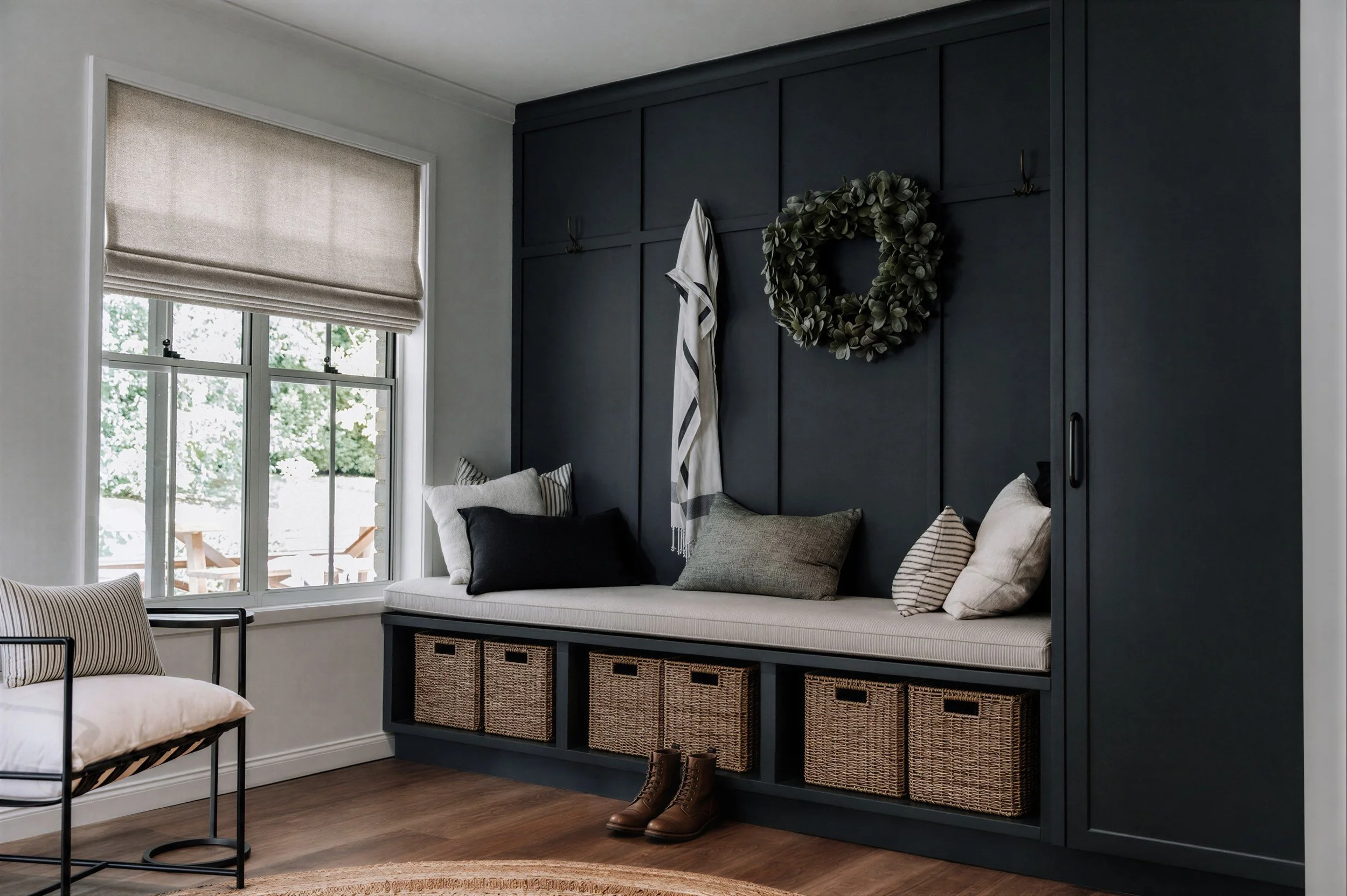 A dark grey green mudroom by Oak & Olive Homes with wainscotting on the wall and a cushioned bench with wicker baskets below for storage