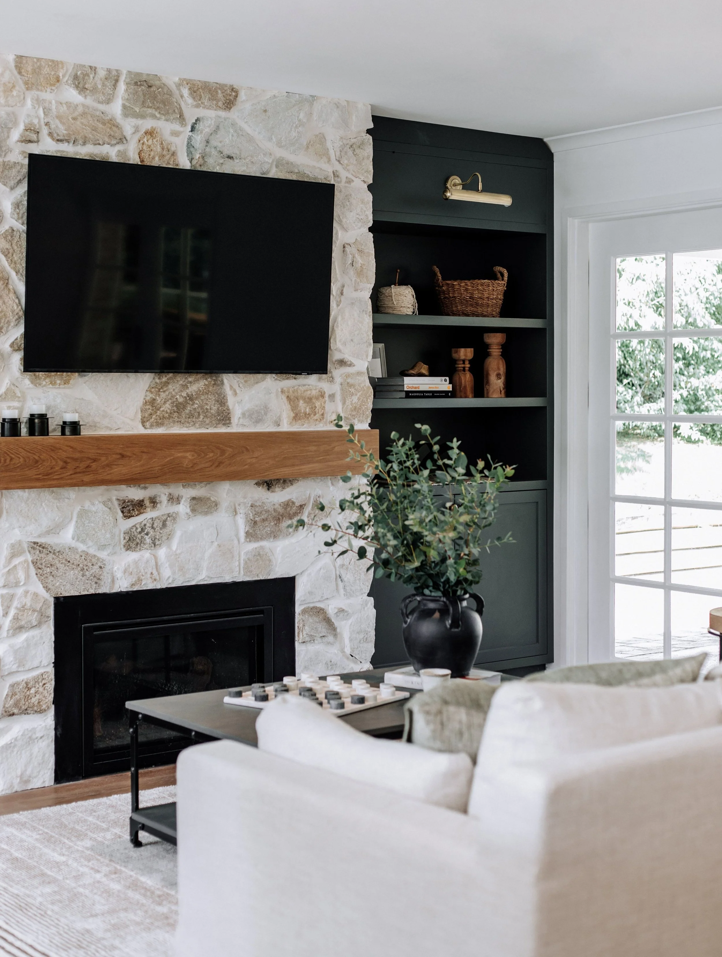 A cozy living room by Oak & Olive Homes featuring a stone clad fireplace with wood mantle and TV above, white French doors leading to the patio, creating a warm and inviting atmosphere