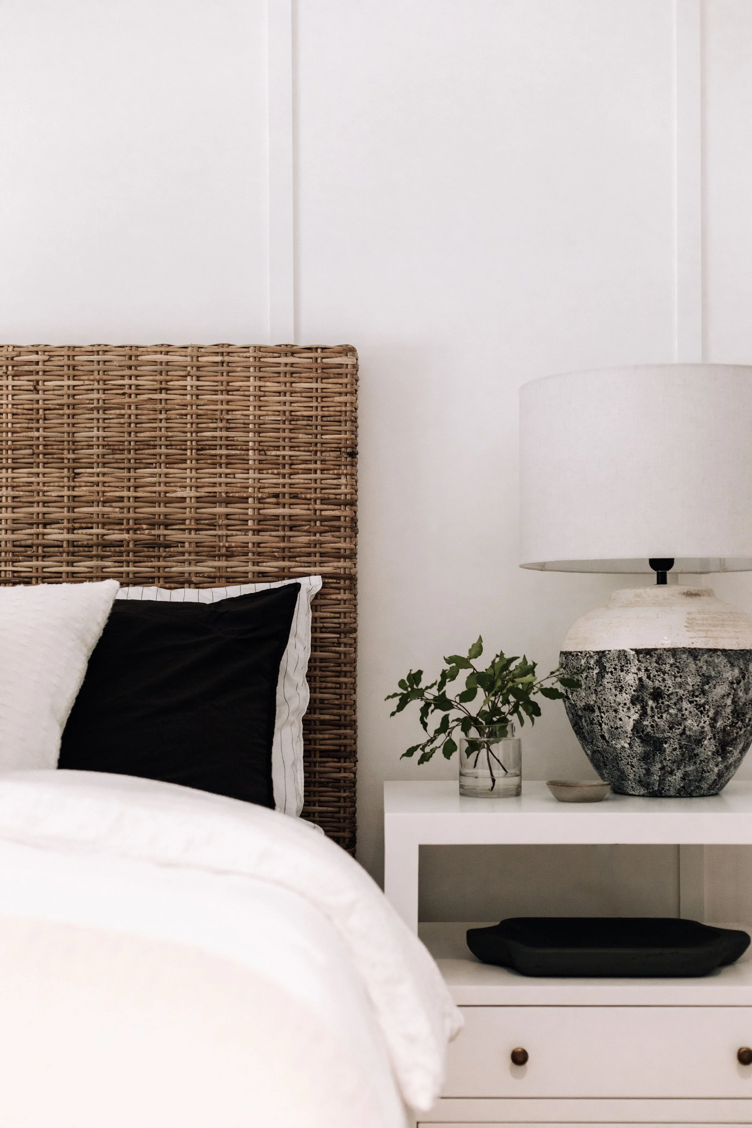 Cozy bedroom vignette featuring a woven rattan headboard, white and black bedding, and a modern white nightstand. Topped with a textured ceramic lamp, glass vase with greenery, and minimal decorative tray for a natural, serene look.