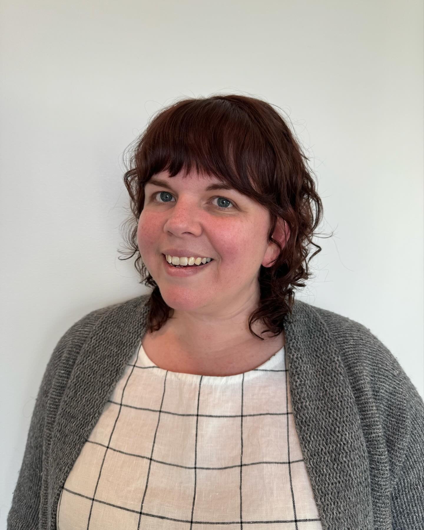 A woman with short, curly, reddish-brown hair, blue eyes, and a light complexion, smiling against a plain white background, wearing a beige top with a black grid pattern and a gray knit cardigan. Haircut by Tiffany Shuck of Rooted Ritual Salon, forme