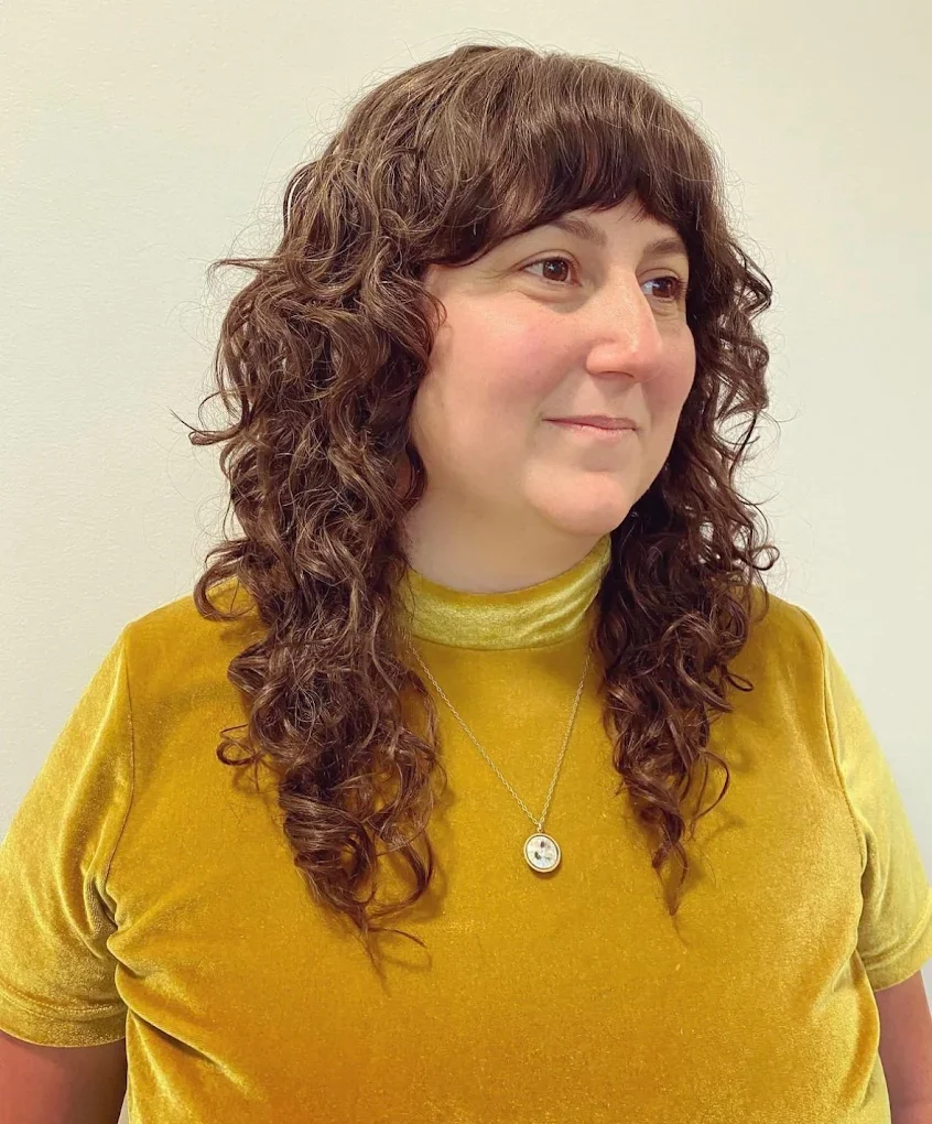A woman with curly brown hair wearing a mustard yellow velvet top and a necklace, standing against a plain light-colored wall.
Haircut by Tiffany Shuck of Rooted Ritual Salon, formerly Hare Salon.