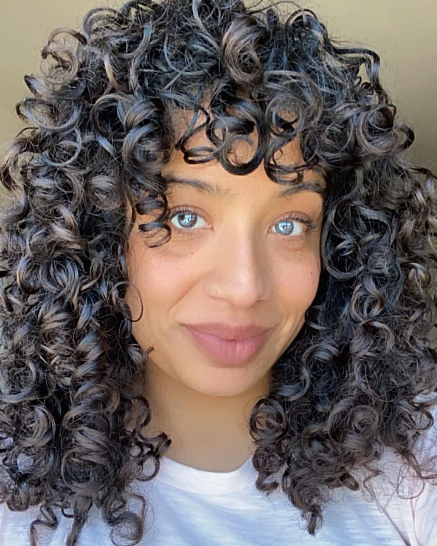 A young woman with curly dark hair and bright blue eyes smiling softly. Haircut by Tiffany Shuck of Rooted Ritual Salon, formerly Hare Salon.