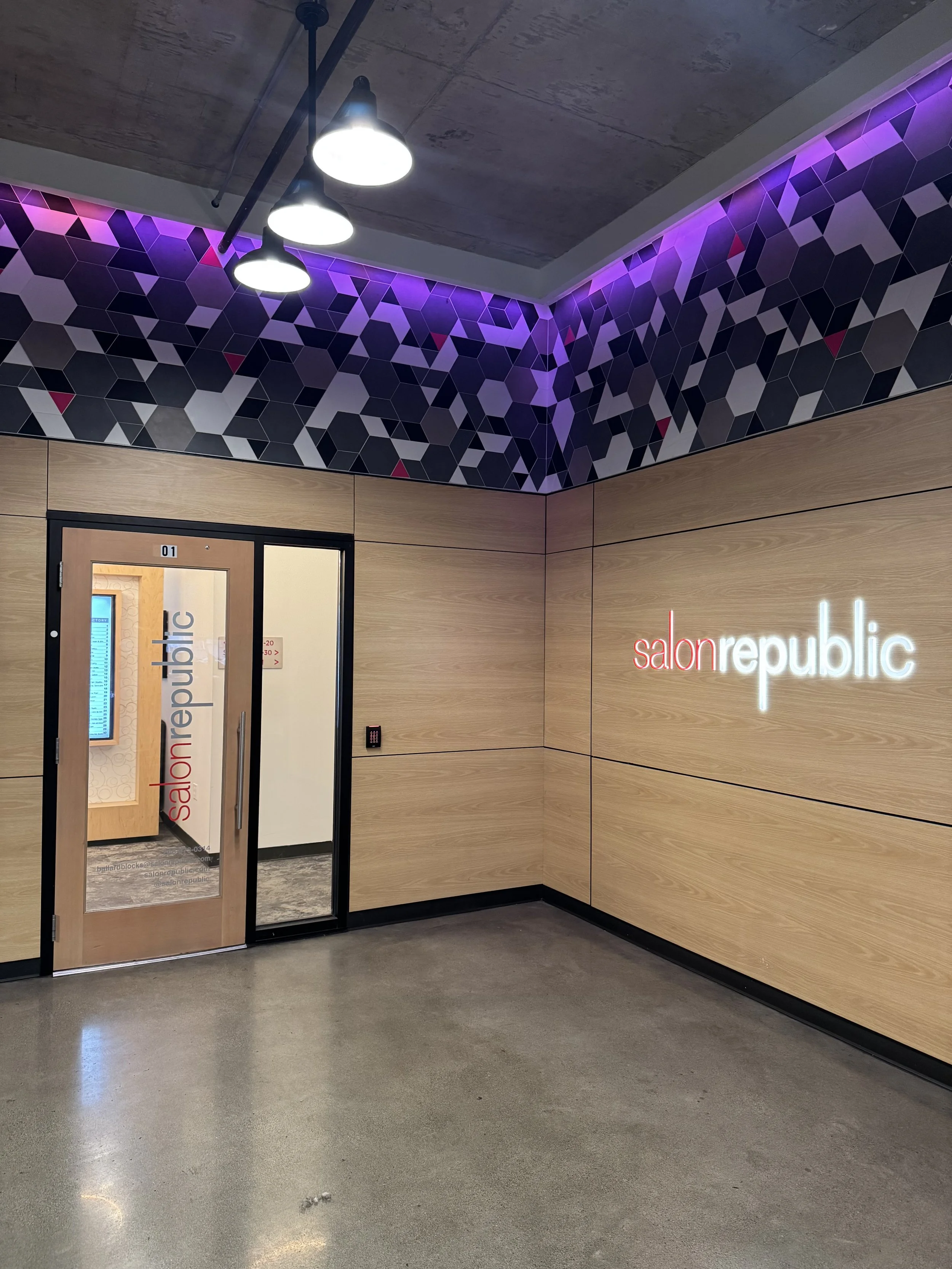 Interior of a modern hair salon with a wooden wall and a neon sign that reads 'salonrepublic'. The ceiling has three hanging lights and purple geometric patterned wallpaper near the ceiling. The entrance has a glass door with the salon's name.