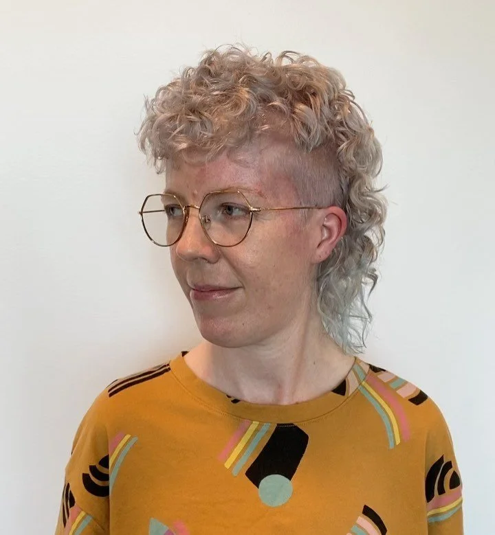 Person with light skin and blond curly hair, wearing round glasses and a yellow shirt with colorful abstract patterns, looking to her left against a plain white background. Haircut by Tiffany Shuck of Rooted Ritual Salon, formerly Hare Salon.