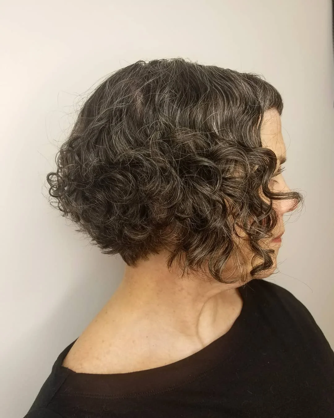 Side view of a woman with short, curly brown hair wearing a black top, standing against a plain light background. Haircut by Tiffany Shuck of Rooted Ritual Salon, formerly Hare Salon.