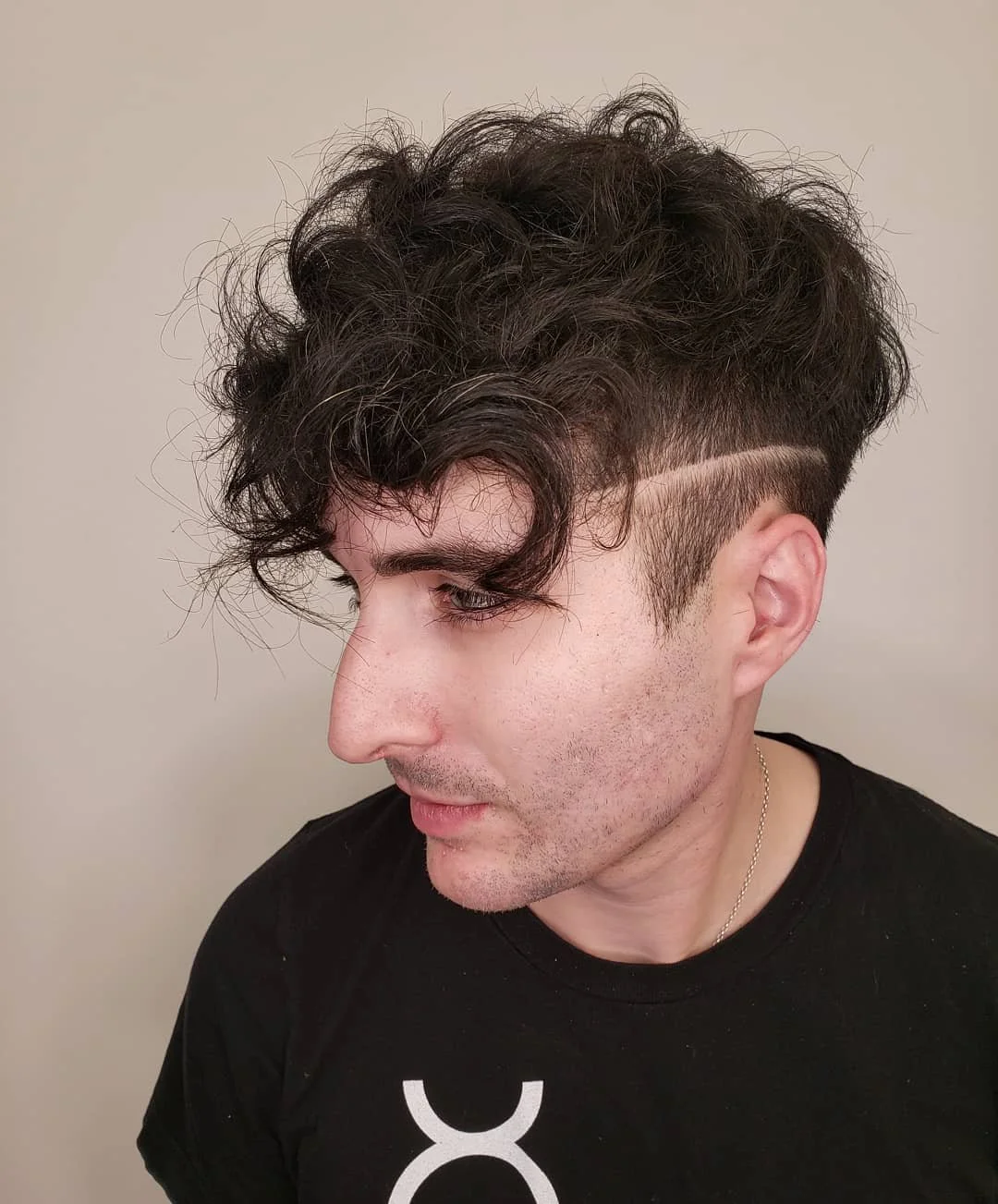 A young person with dark curly hair and fade haircut, featuring a shaved line design, wearing a black shirt with a white logo. Haircut by Tiffany Shuck of Rooted Ritual Salon, formerly Hare Salon.