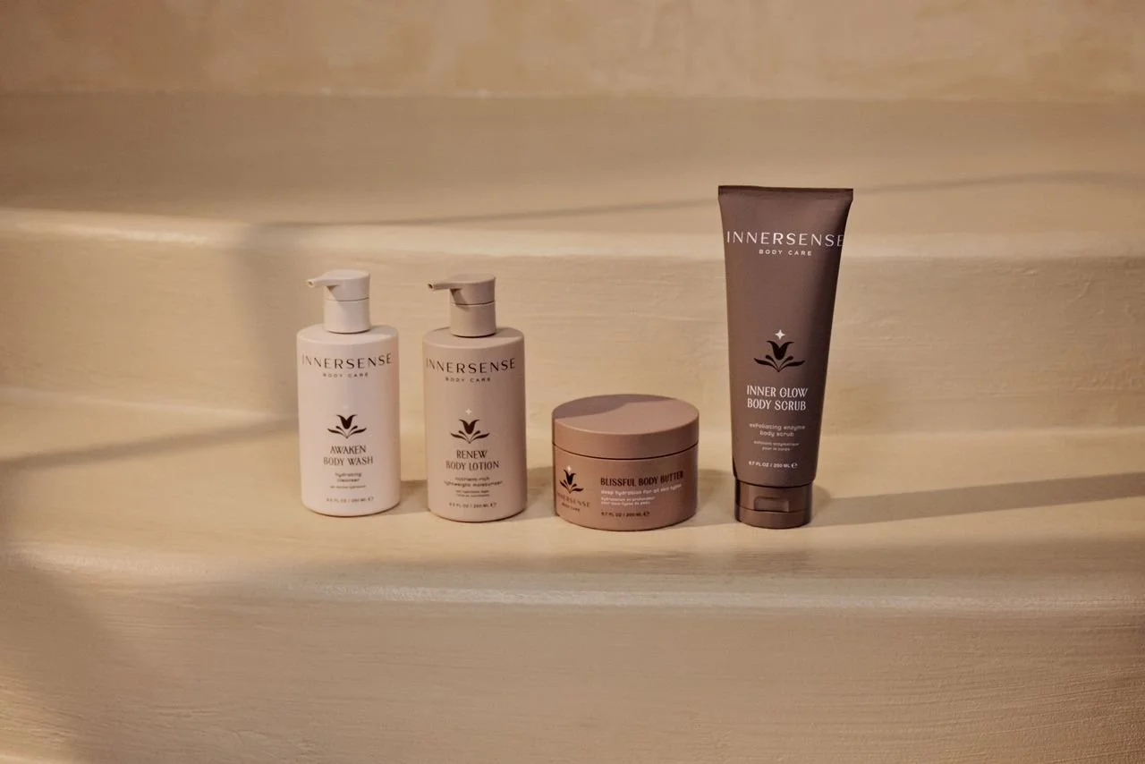 Four skincare products from InnerSense arranged on a wooden surface: Awaken Body Wash, Renew Body Lotion, Blissful Body Butter, and Inner Glow Body Scrub. Innersense sold at Rooted Ritual Salon.