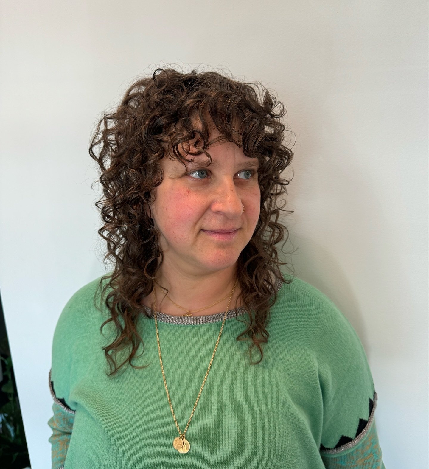 A woman with curly brown hair and blue eyes wearing a green sweater and gold necklaces, standing against a plain white wall. Haircut by Tiffany Shuck of Rooted Ritual Salon, formerly Hare Salon.