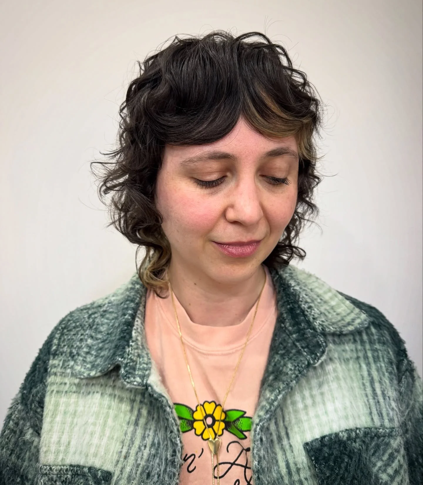 A woman with curly dark hair and blonde highlights, wearing a pink shirt with a floral design, a gray and green checkered jacket, and a gold necklace with a yellow flower pendant, looking down with a gentle smile. Haircut by Tiffany Shuck of Rooted R