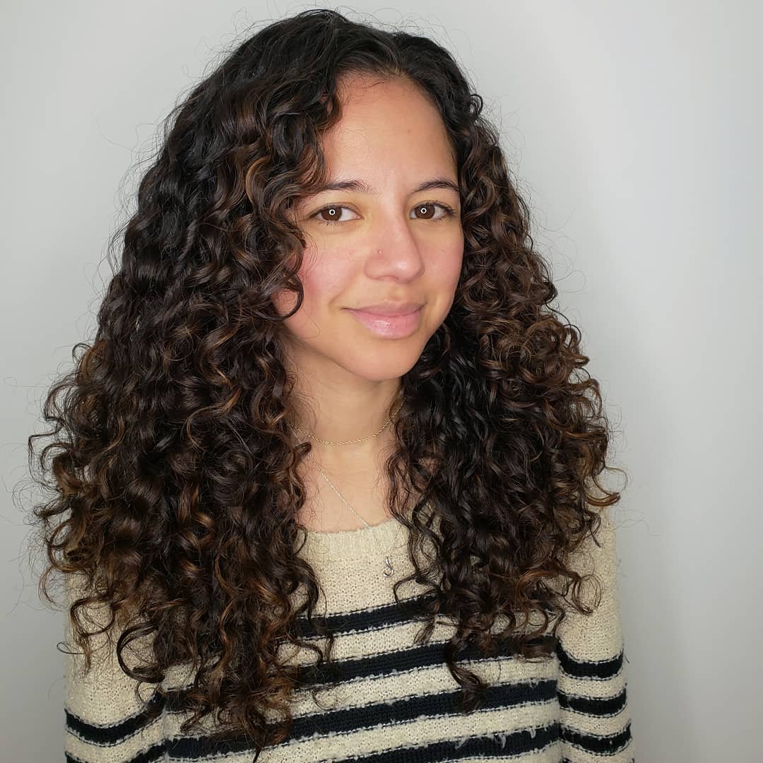 A woman with long, curly dark brown hair, wearing a beige and black striped sweater, smiling softly against a plain white background. Haircut by Tiffany Shuck of Rooted Ritual Salon, formerly Hare Salon.