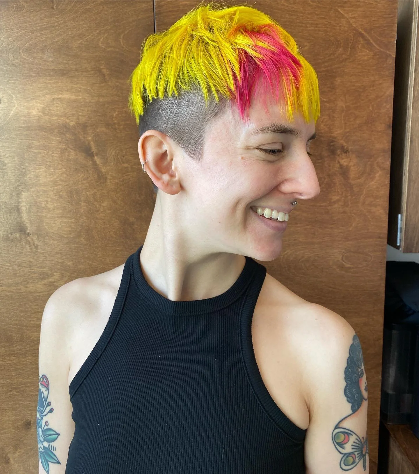 Profile of a woman with a bright yellow and pink pixie haircut, wearing a black sleeveless top, and displaying tattoos on her arm and shoulder, standing against a wooden background. Haircut by Tiffany Shuck of Rooted Ritual Salon, formerly Hare Salon