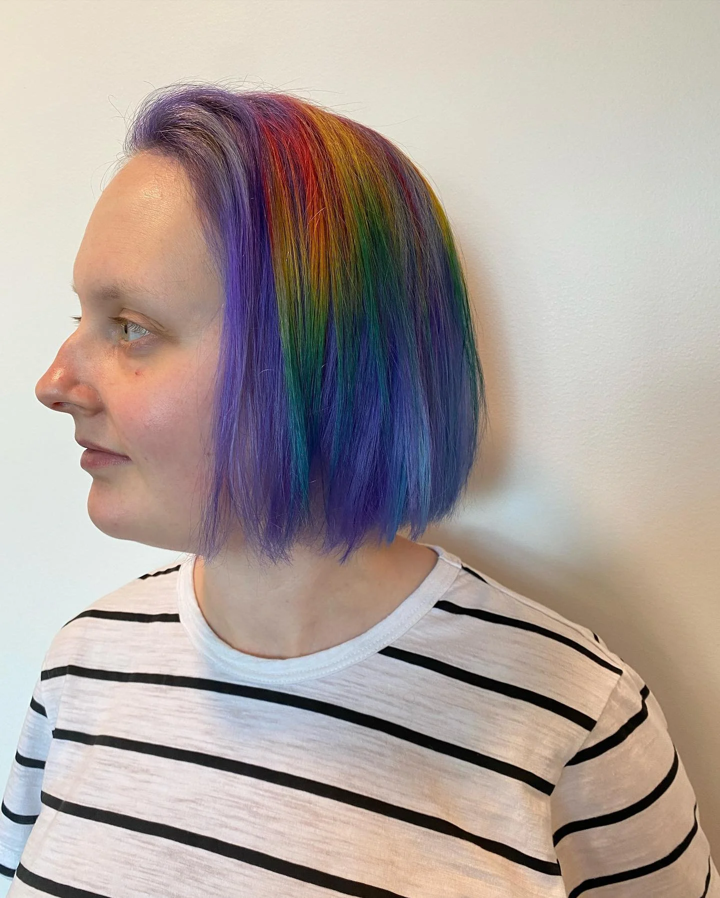Woman with short hair dyed in rainbow colors including purple, blue, green, yellow, orange, and red, wearing a white shirt with black horizontal stripes, standing against a plain white wall.Haircut by Tiffany Shuck of Rooted Ritual Salon, formerly Ha