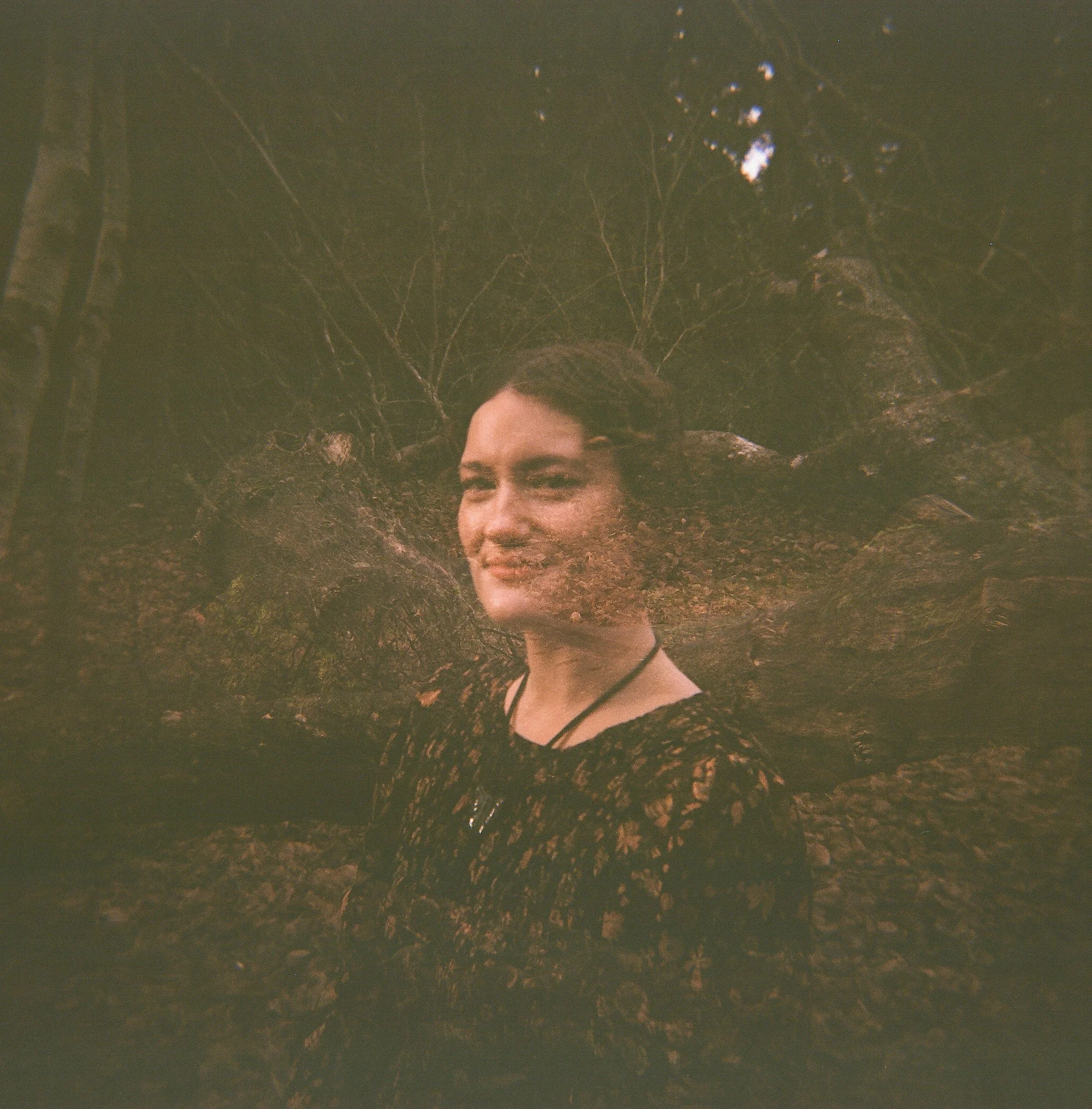 Double exposure photograph of Tiffany Shuck of Rooted Ritual Salon with short dark hair, standing in front of a fallen tree trunk with bare branches in the background.