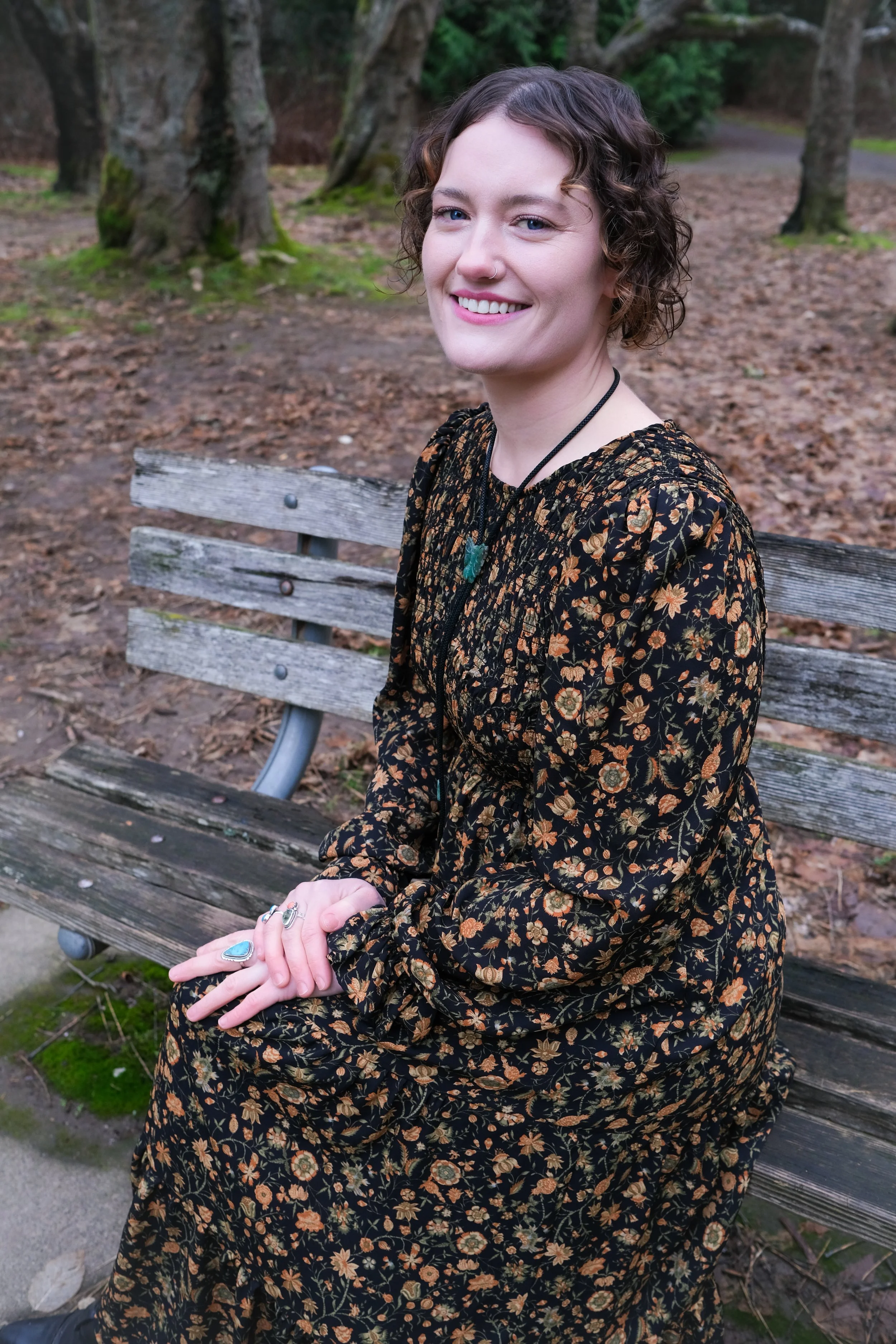 Tiffany Shuck previous co-owner of Hare Salon with short curly brown hair sitting on a wooden park bench in a wooded area, smiling at the camera, wearing a black floral dress with orange and yellow flowers, and accessorized with turquoise jewelry.