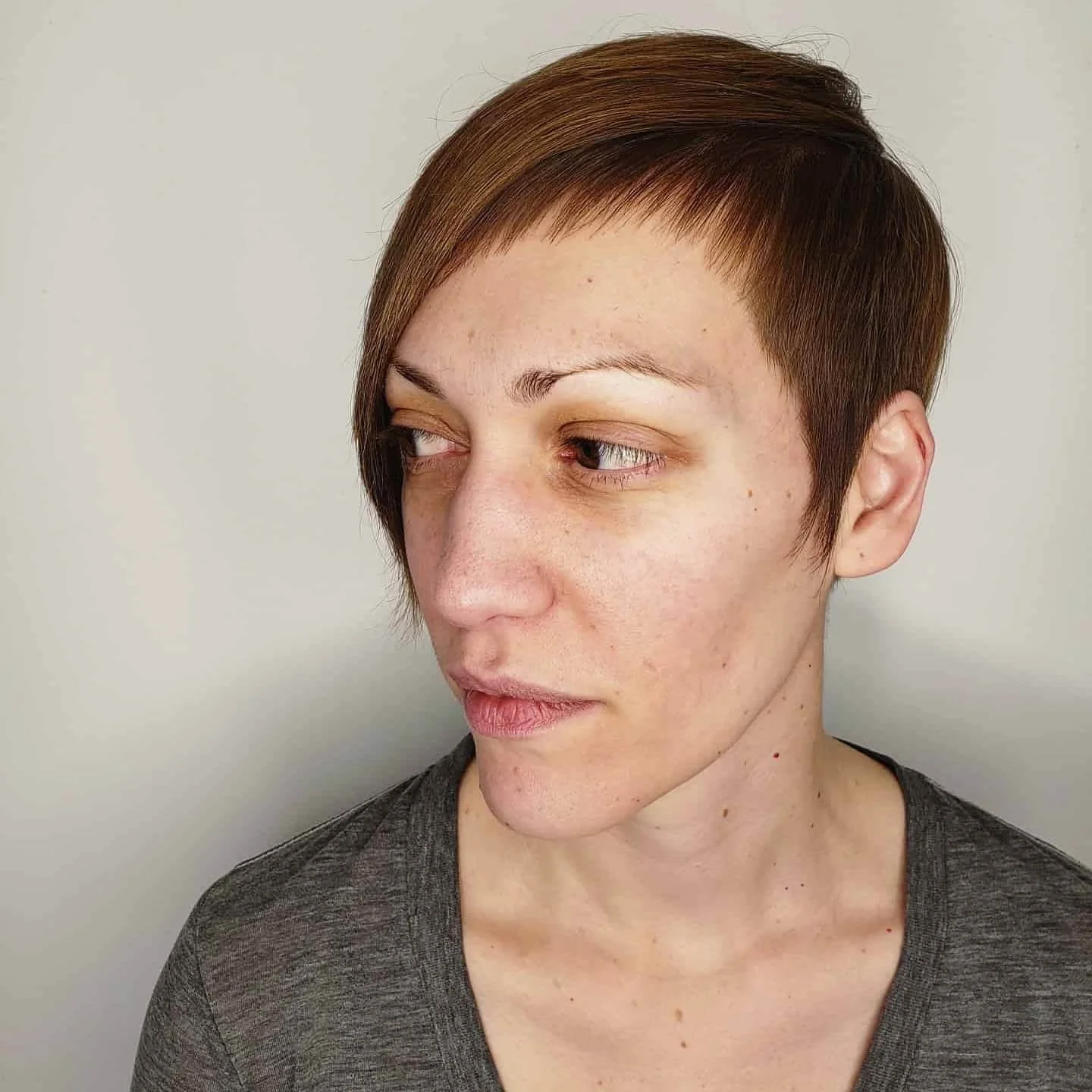 A woman with short brown hair looking to her left, wearing a gray shirt against a plain white background. Haircut by Tiffany Shuck of Rooted Ritual Salon, formerly Hare Salon.