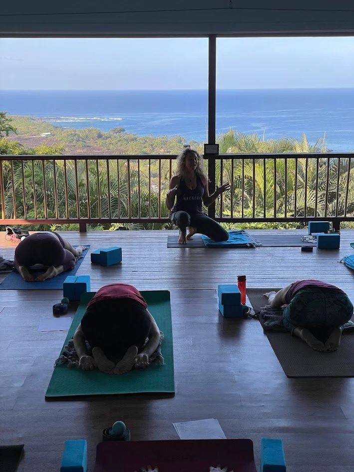 Indoor yoga class with a view of the ocean through large windows, instructors leading students in a seated forward bend pose.