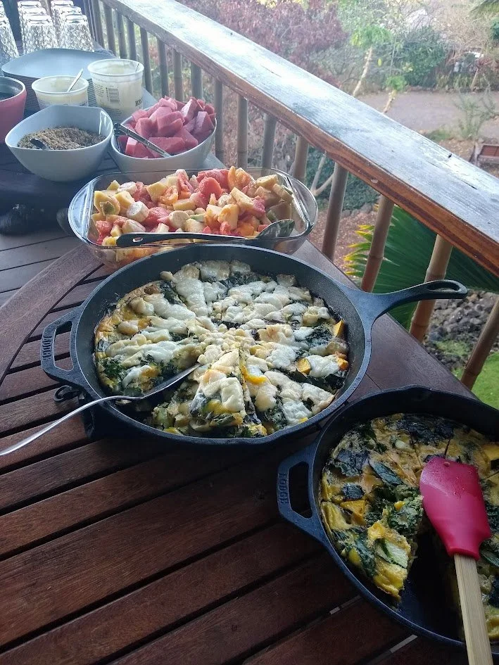 Fried vegetable frittata in a cast iron skillet, with a spatula resting inside. Next to it is a smaller skillet with a vegetable frittata. In the background, a bowl of mixed fruit salad, a bowl of raw pink meat, and bowls of spices and condiments on 