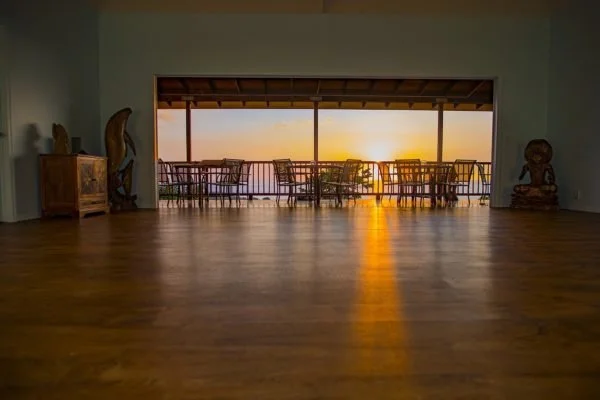 Room with wooden floor and large sliding glass doors leading to a balcony with a sunset view. The balcony has several chairs and a table. Decorative items are on the sides of the room.