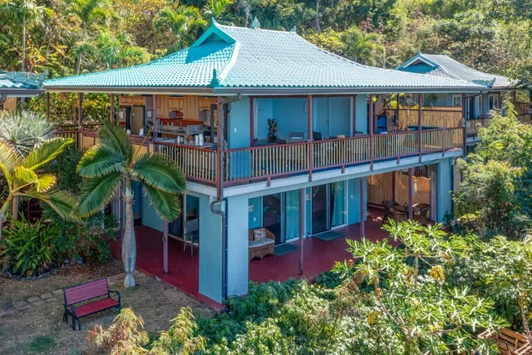 A two-story blue house with a large outdoor balcony, surrounded by lush tropical plants and trees, with a sloped roof and a garden area with a bench in the foreground.